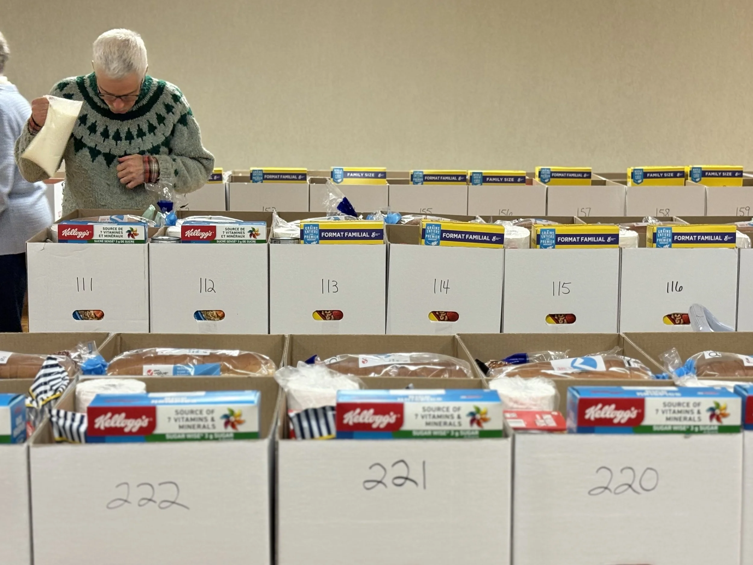 Man with white hair and glasses shopping for groceries, looking into boxes of food items on a table in an indoor setting.