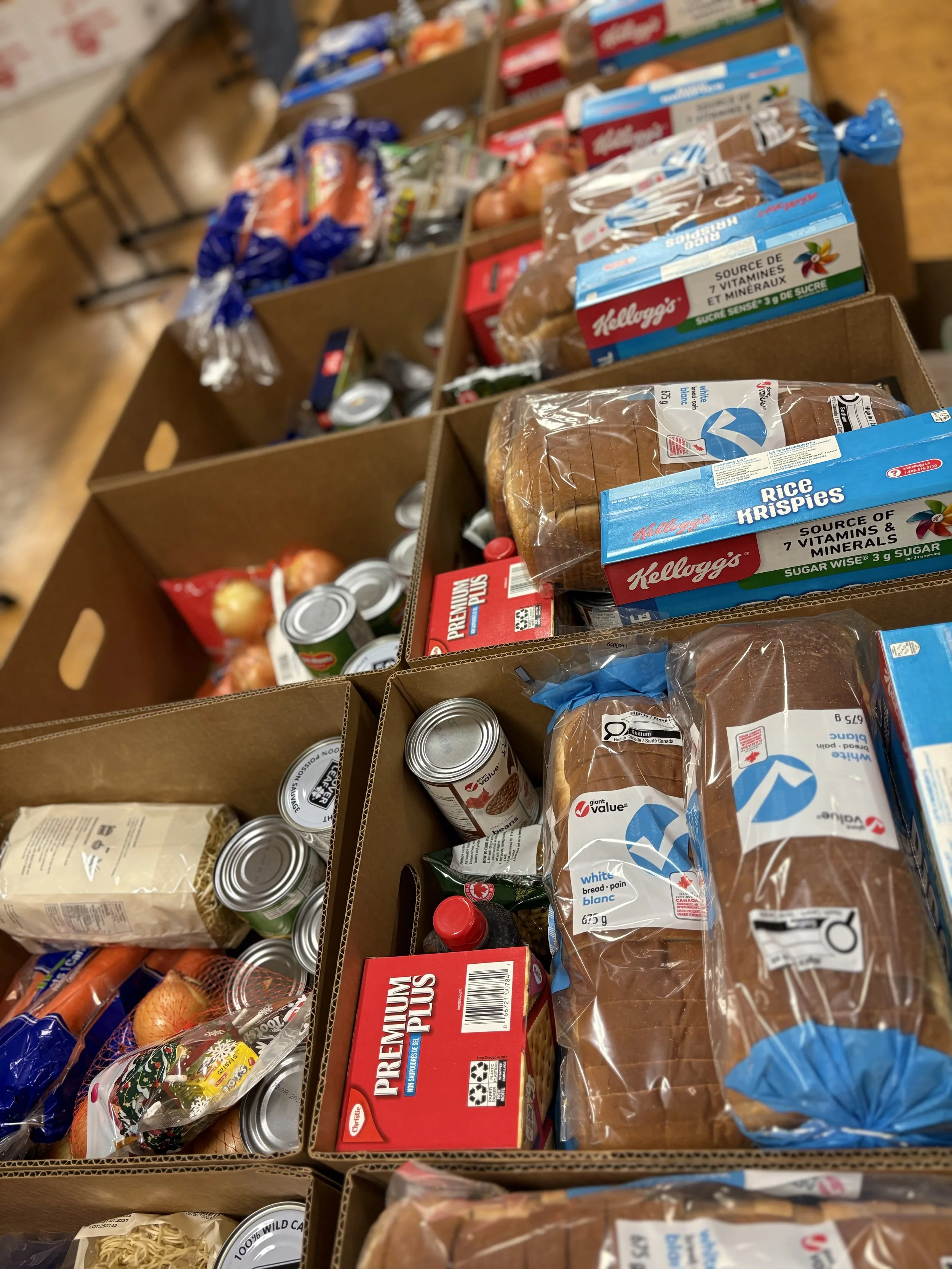 Multiple cardboard boxes filled with groceries such as bread, canned goods, onions, and other packaged food items on a wooden table.
