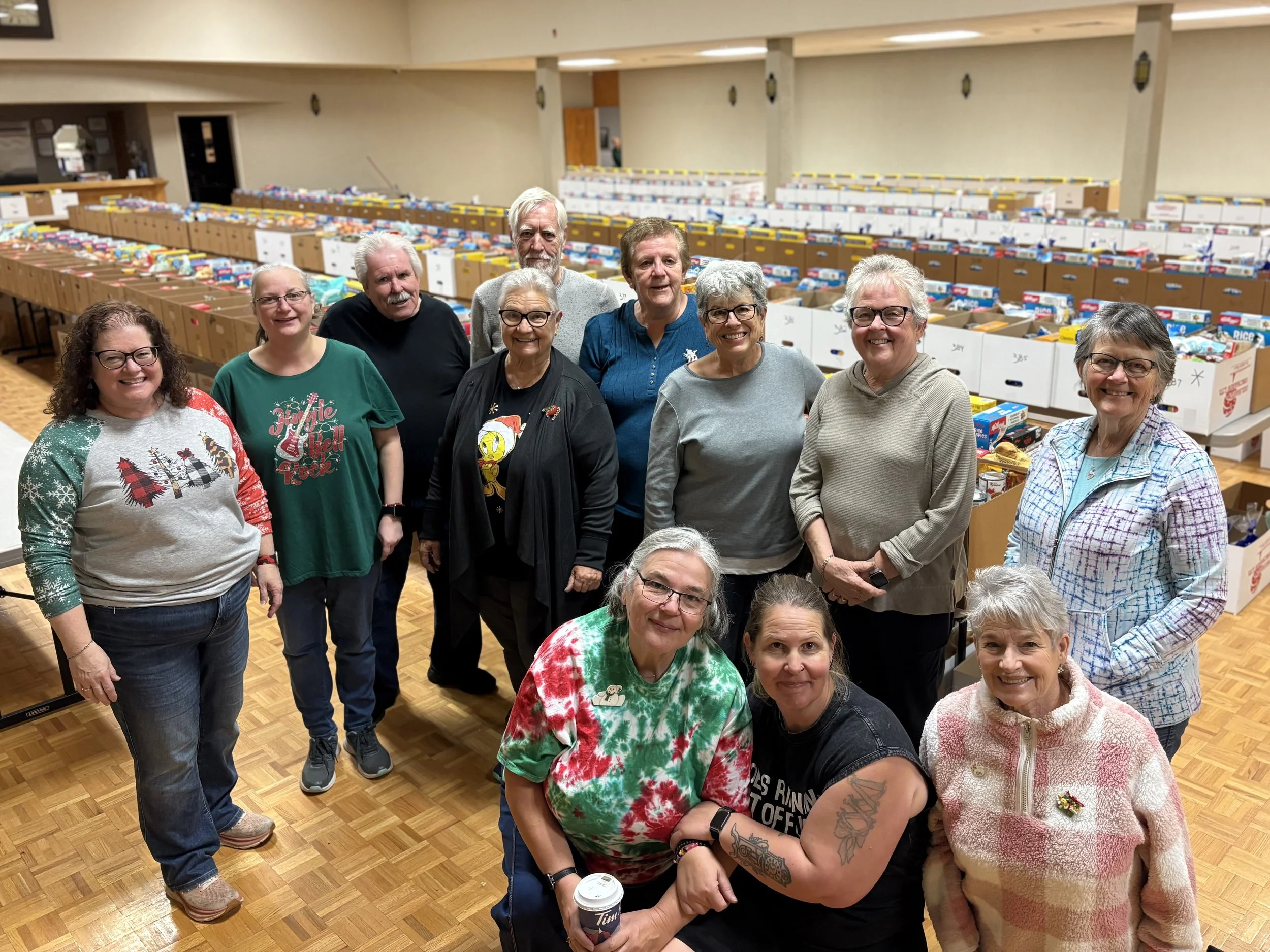 Group of ten smiling volunteers indoors, standing in front of tables filled with boxes, possibly for a charitable event.