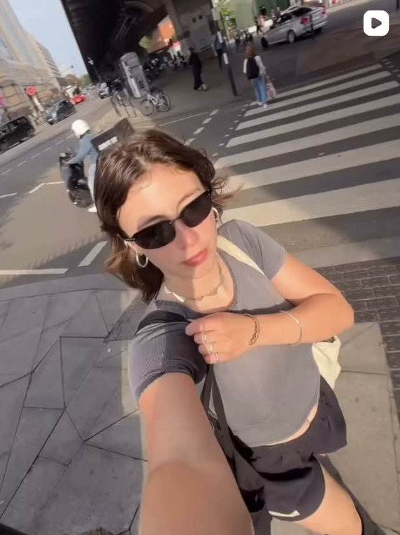 A woman with short brown hair wearing black sunglasses, a gray t-shirt, and earrings, taking a selfie on a city street with crosswalk and traffic in the background.