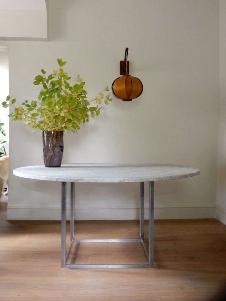 Round white marble table with metal legs, a purple vase with green leafy branches, a wooden wall-mounted lamp, and a partial view of a dog tail in a room with wooden floors and light-colored walls.