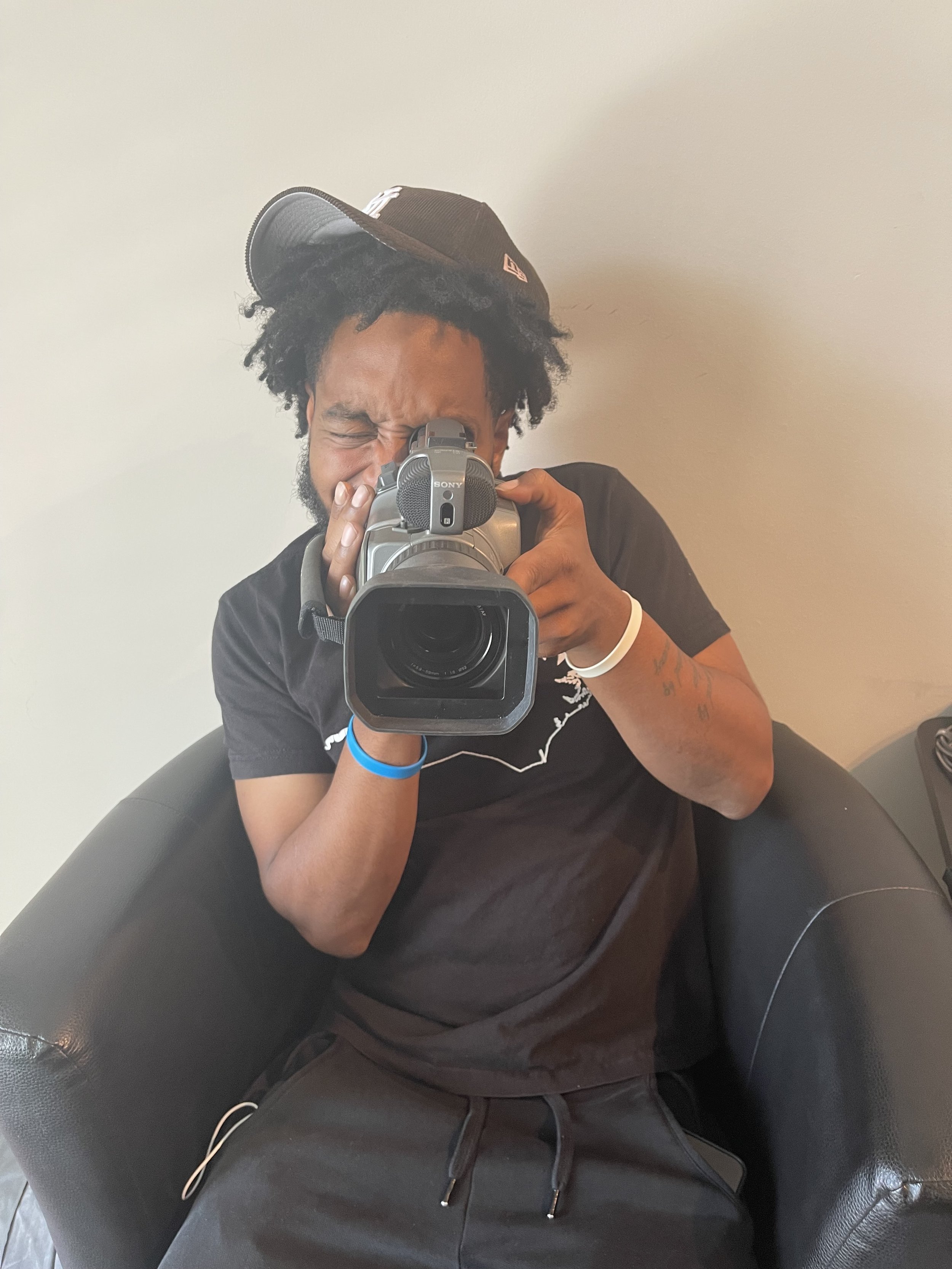 A man with black curly hair and a beard taking a selfie with a video camera in front of a plain beige wall. He wears a black t-shirt, a white wristband, and a black cap tilted upside down under the camera.
