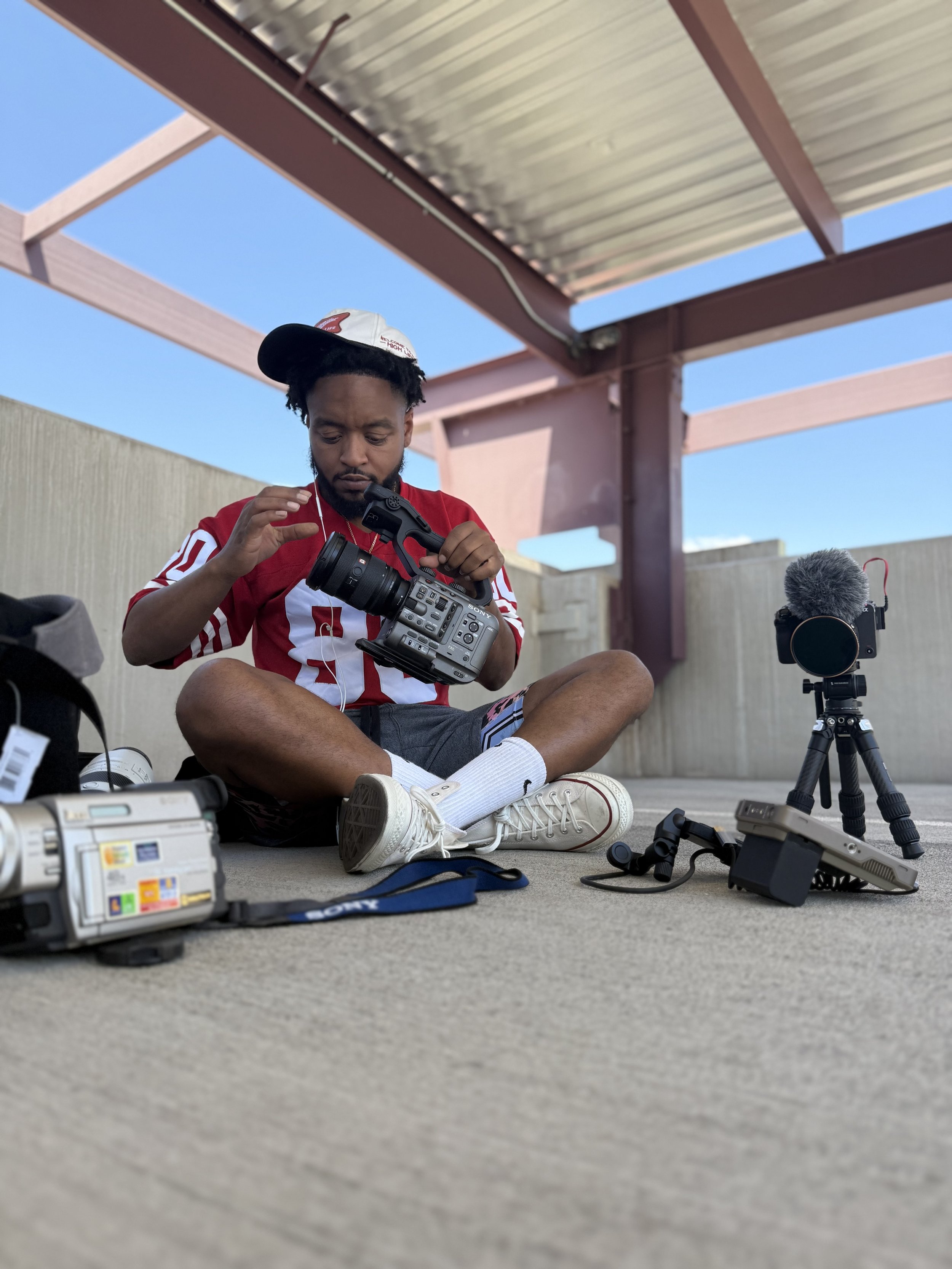 A man sitting cross-legged on the ground, adjusting a professional video camera, with various camera equipment scattered around him under a metal roof structure.