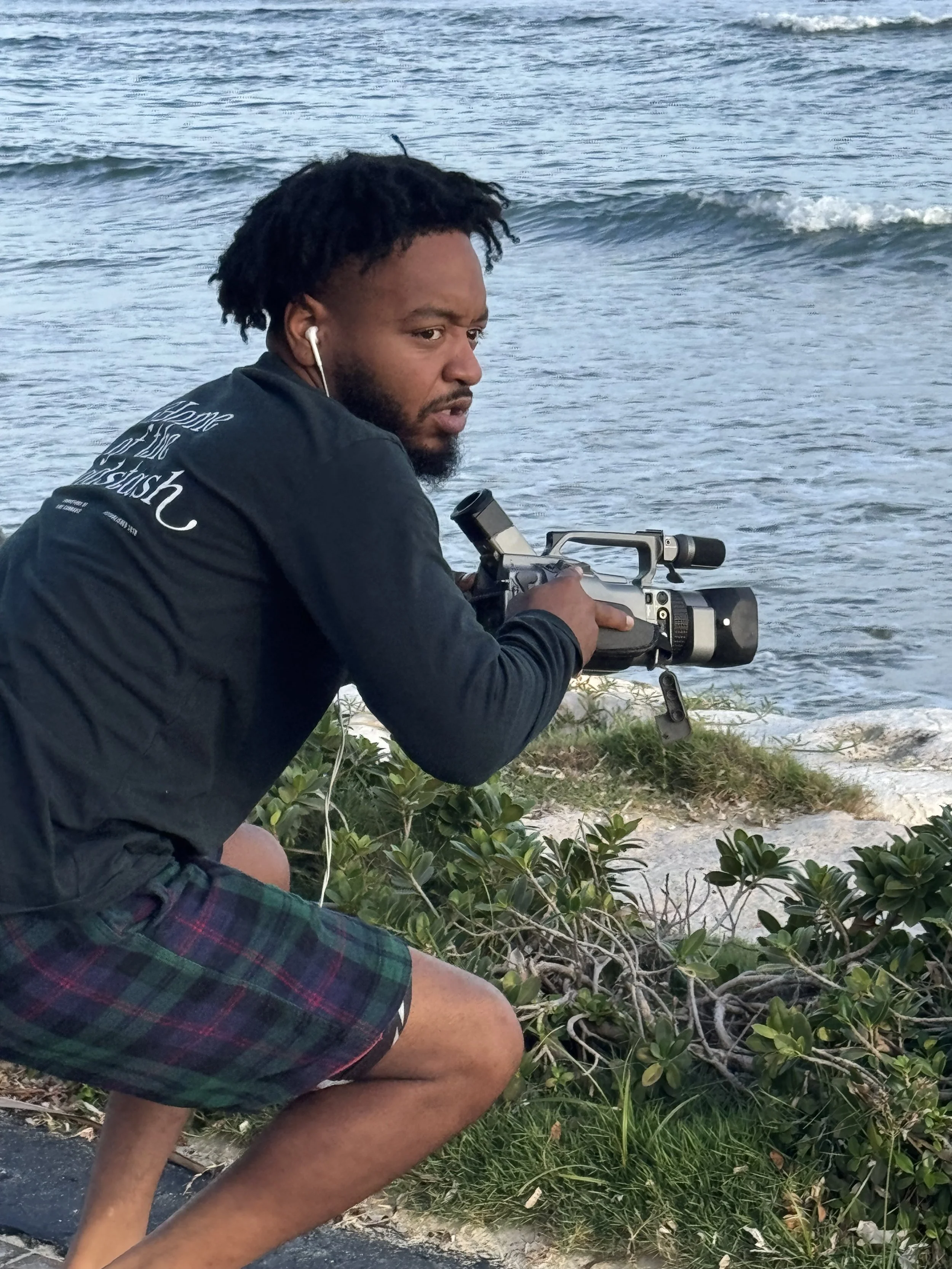 A man with dark curly hair and a beard crouches near the edge of a body of water, holding a camera with a large lens. He is wearing a dark jacket, plaid shorts, and white earphones. Green plants and rocks are visible on the shore, with small waves in the water in the background.