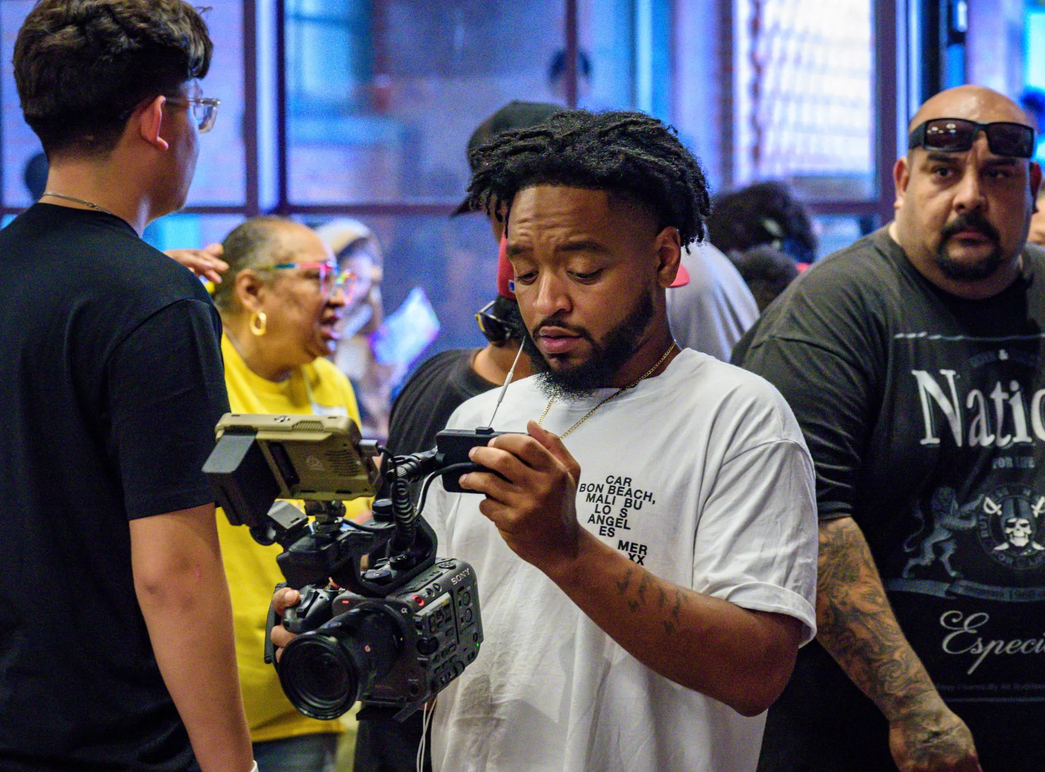 A group of people, including a young man with dreadlocks and a white t-shirt, looking at a camera in an indoor setting with large windows and other individuals in the background.