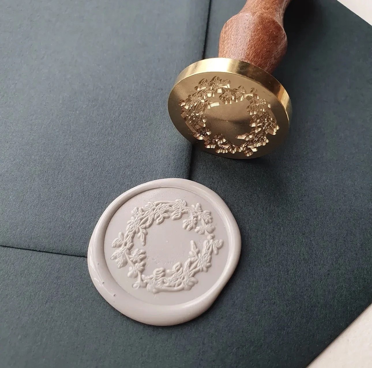 A rubber stamp with a floral wreath design and a white wax seal with the same floral wreath pattern, placed on a dark teal surface.