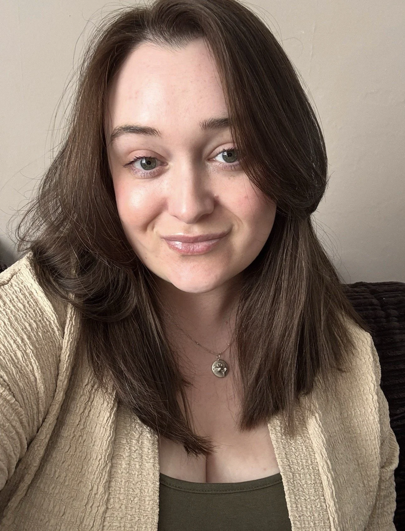 Close-up of a woman with long brown hair, green eyes, wearing a beige textured jacket and a silver necklace with a round pendant, smiling gently at the camera.