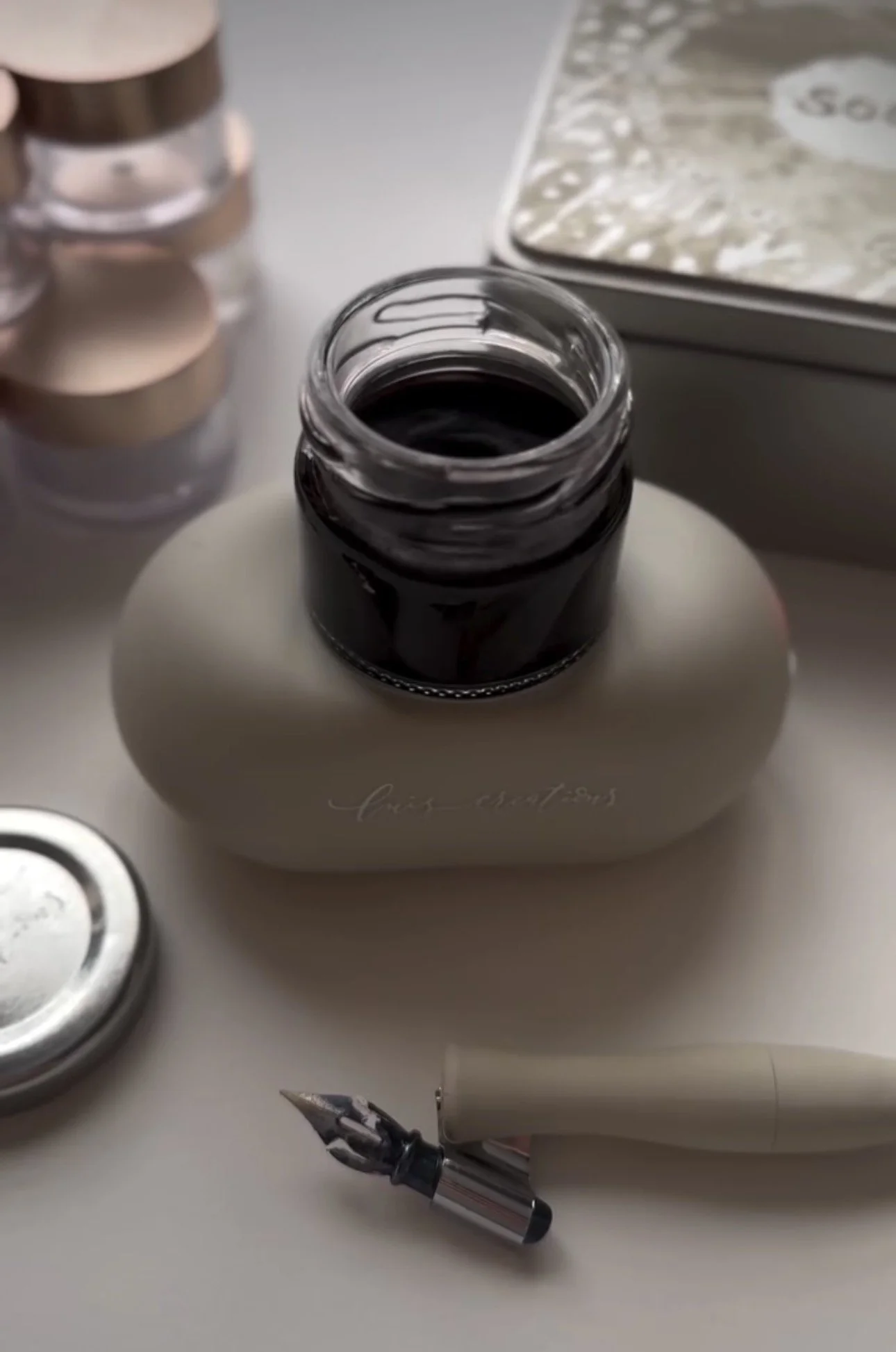 An open jar of black ink, a fountain pen nib, and a collection of copper-colored coins on a white surface. The scene appears to be a workspace or desk setup.