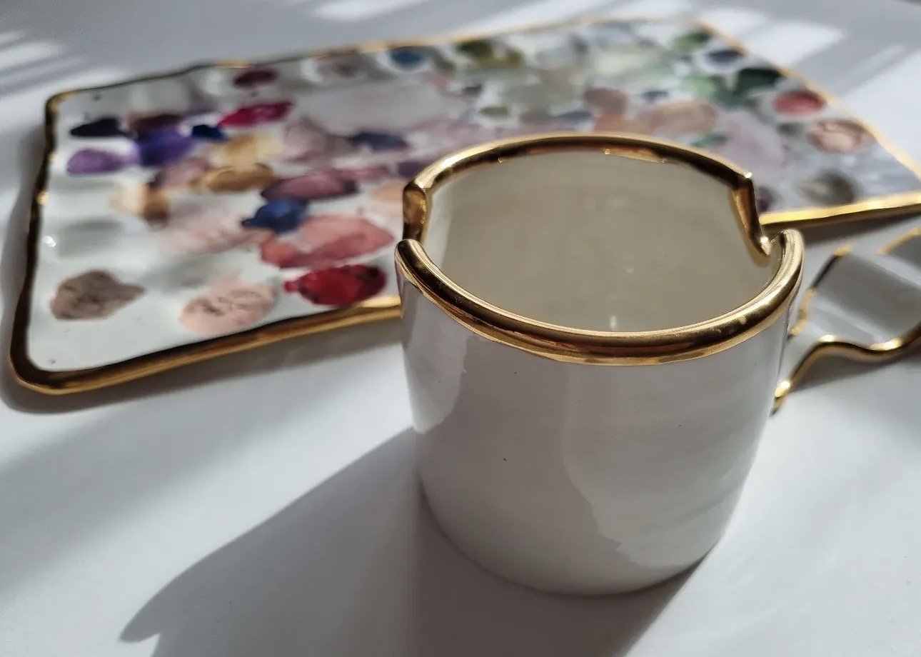 A white ceramic cup with gold trim and a handle, filled with a hot beverage, placed on a white surface. Behind it are two floral-patterned porcelain trays with gold edges.