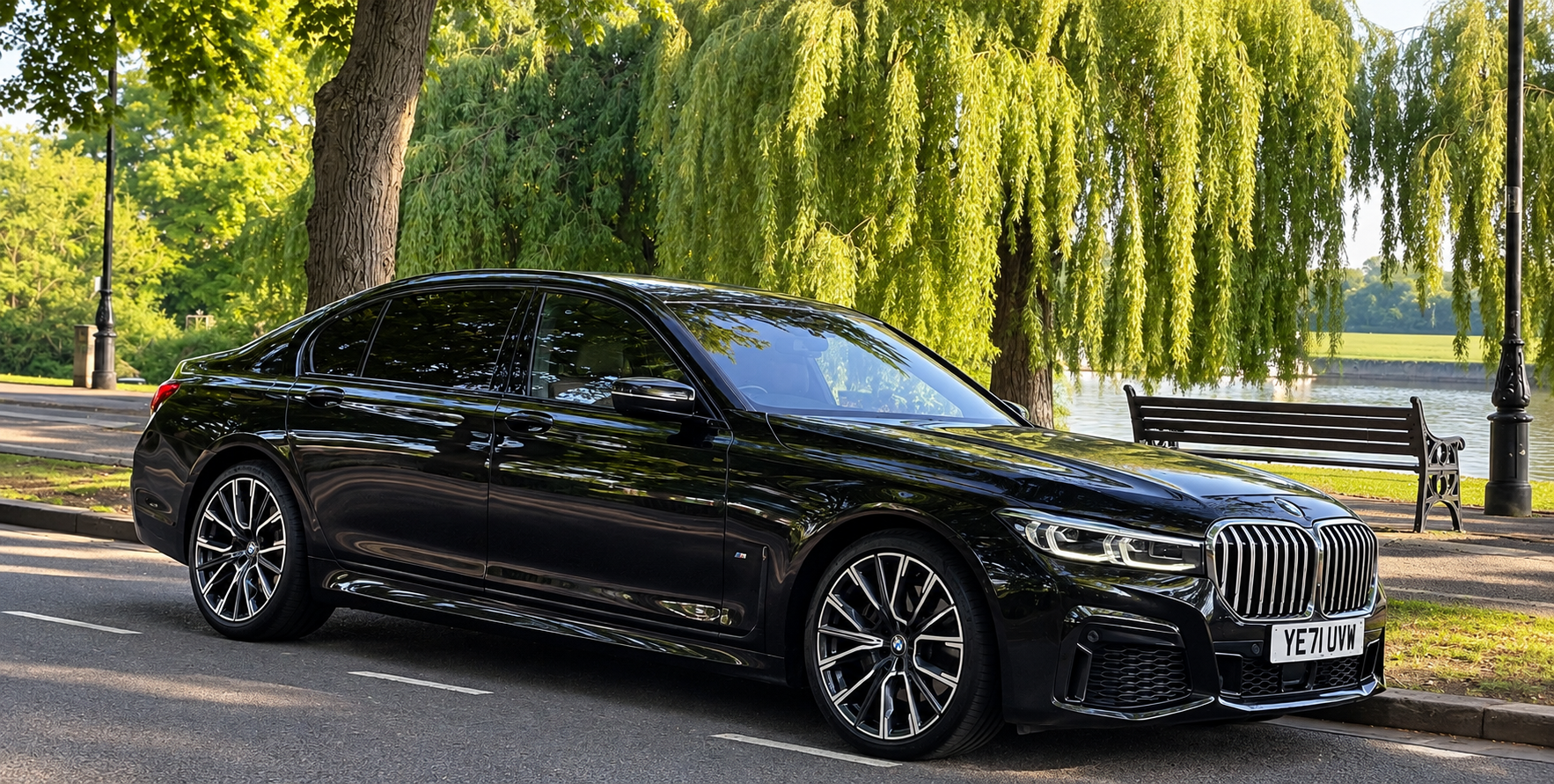 Black BMW 7 Series parked beside a landscaped park and lake.