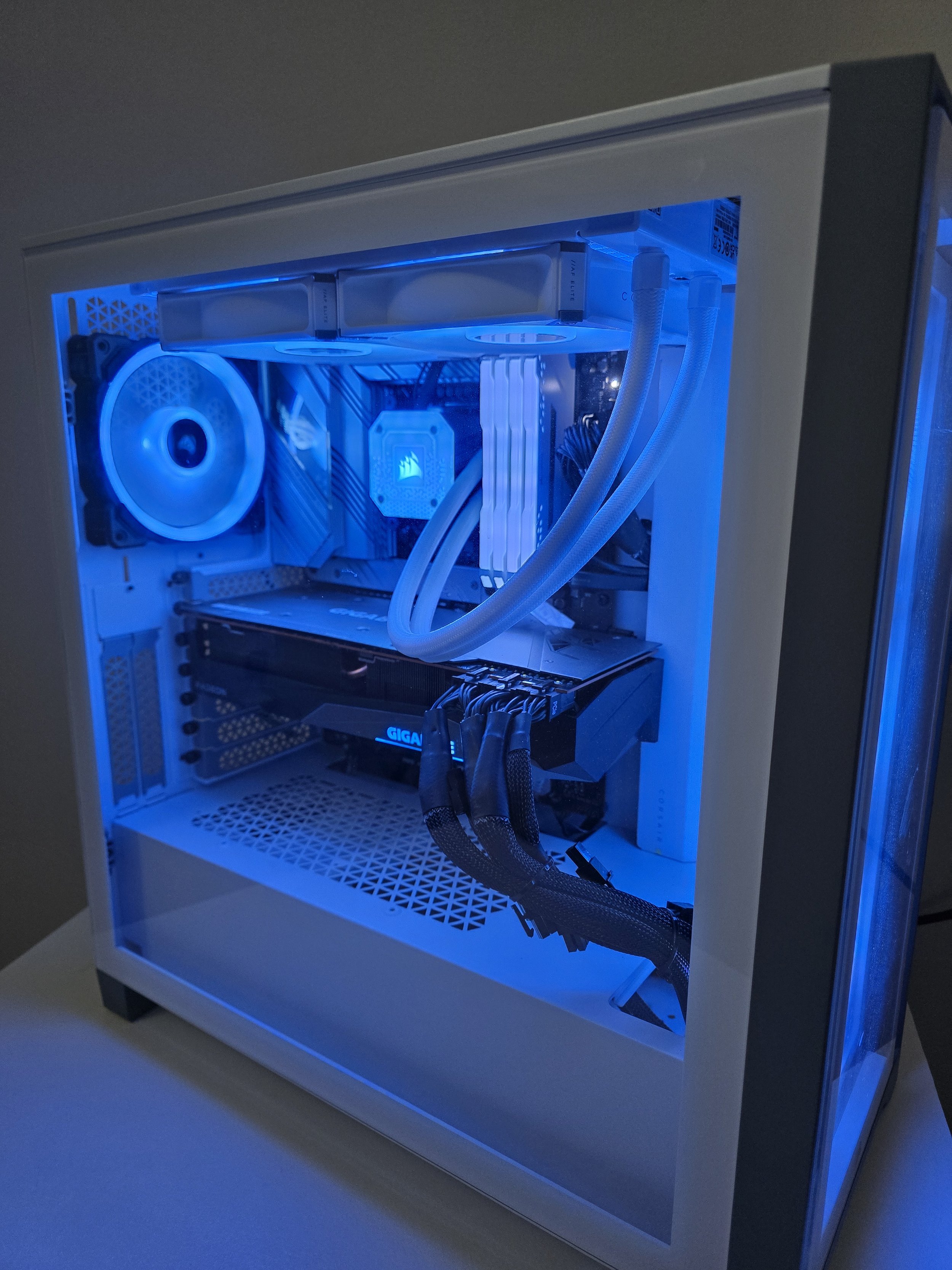 A white computer case with a glass side panel revealing internal components illuminated by blue LED lighting. Inside are a cooling fan, a liquid cooling radiator, cables, and a graphics card.