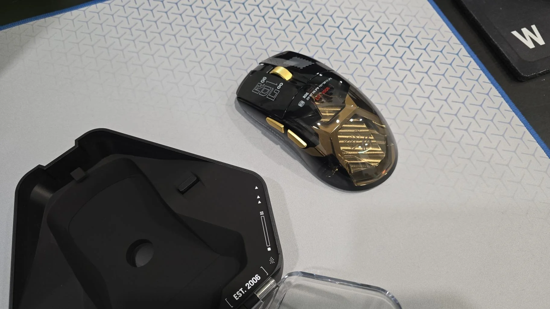 A black and gold gaming mouse on a white textured surface with a plastic case and a black object with a 'W' on it nearby.