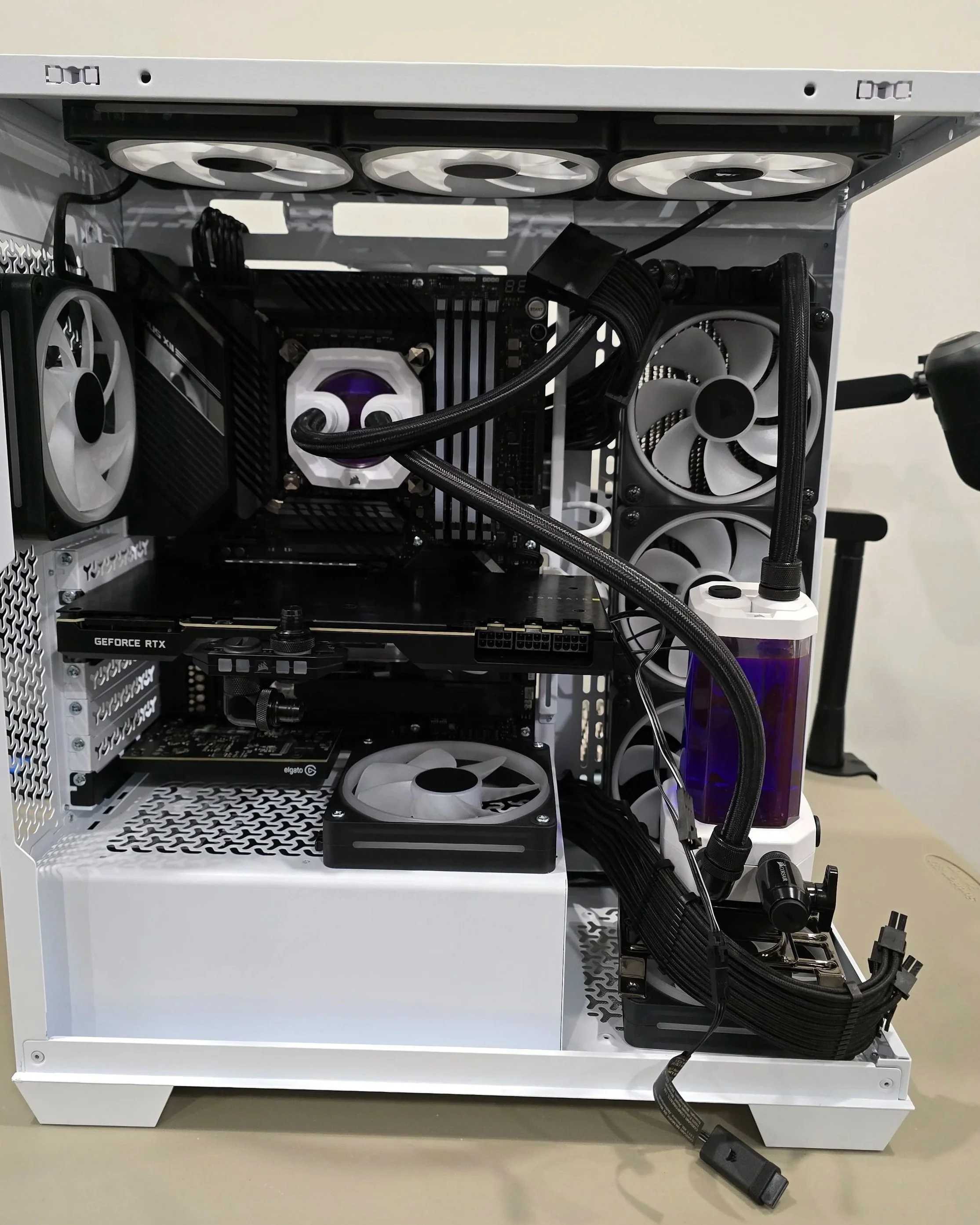 A partially assembled white computer case with black and white fans, a graphics card, a CPU cooler with liquid cooling tubes, power supply, and cables inside, placed on a light-colored surface.