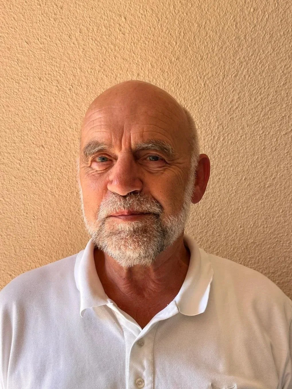 Ein älterer Mann mit grauem Bart und blauen Augen trägt ein weißes Polohemd und steht vor einer beige gekachelten Wand.