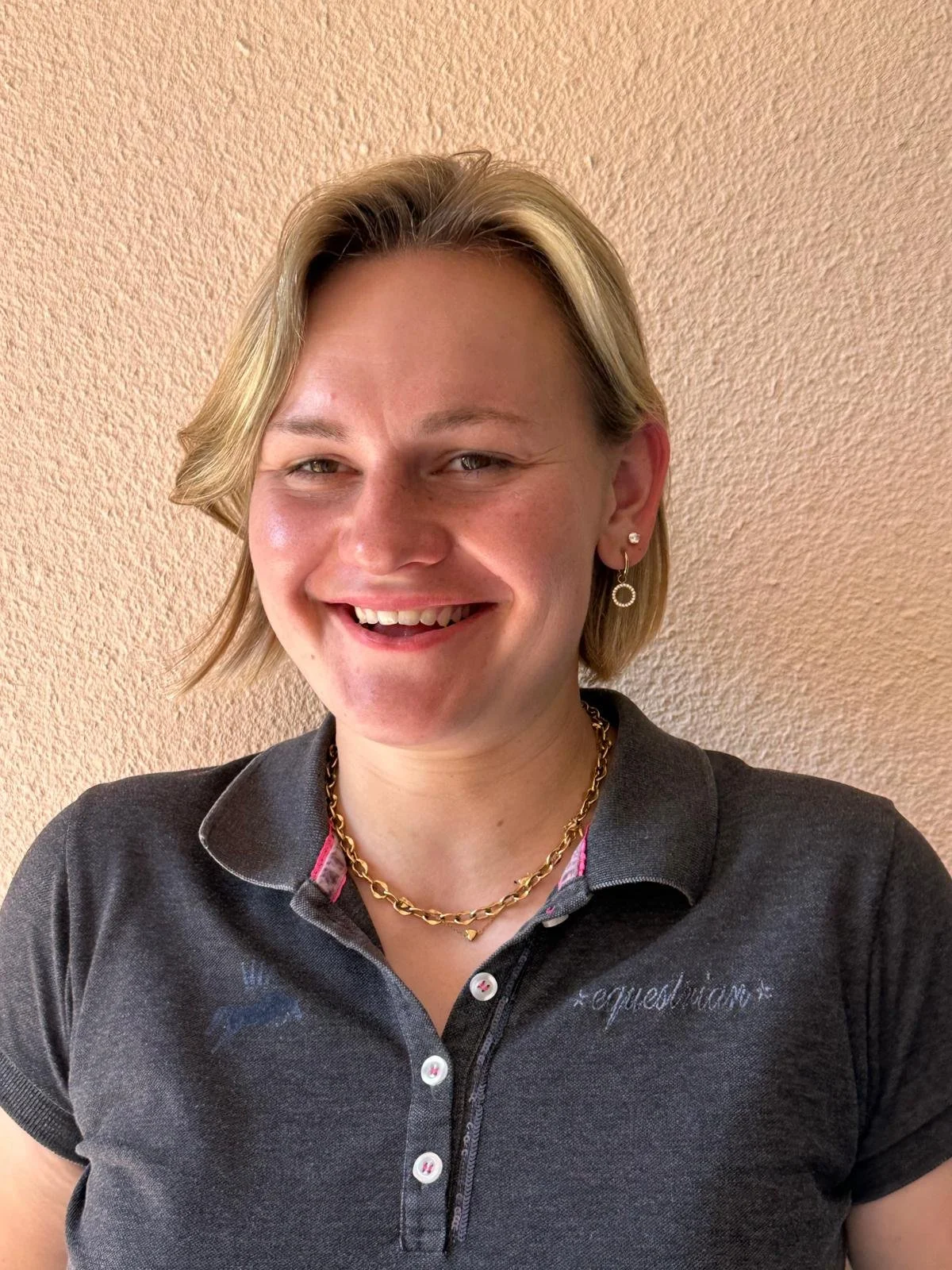Junge Frau mit Kurzhaarfrisur, trägt eine schwarze Piqueshirt und goldene Halskette, lächelt vor hellbraunem Wand.