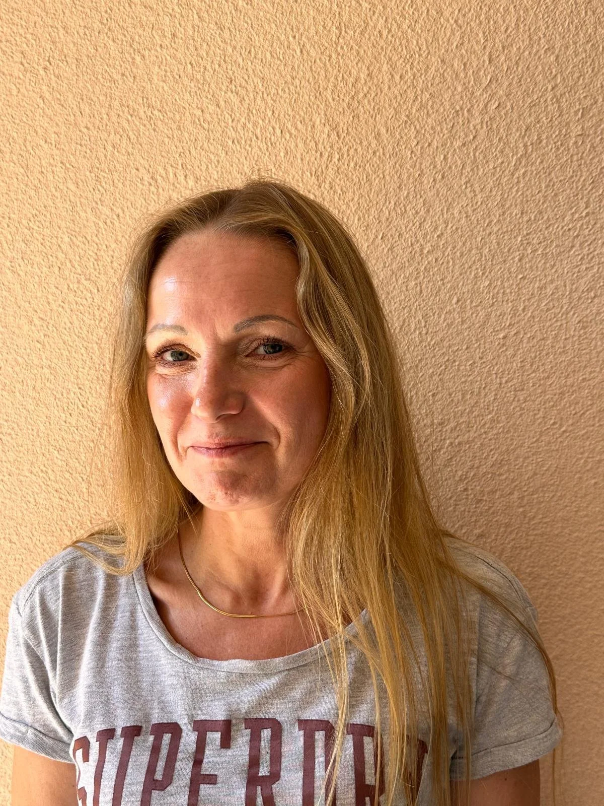 Eine lächelnde Frau mit langen blonden Haaren vor einer peachy-orange Wand.