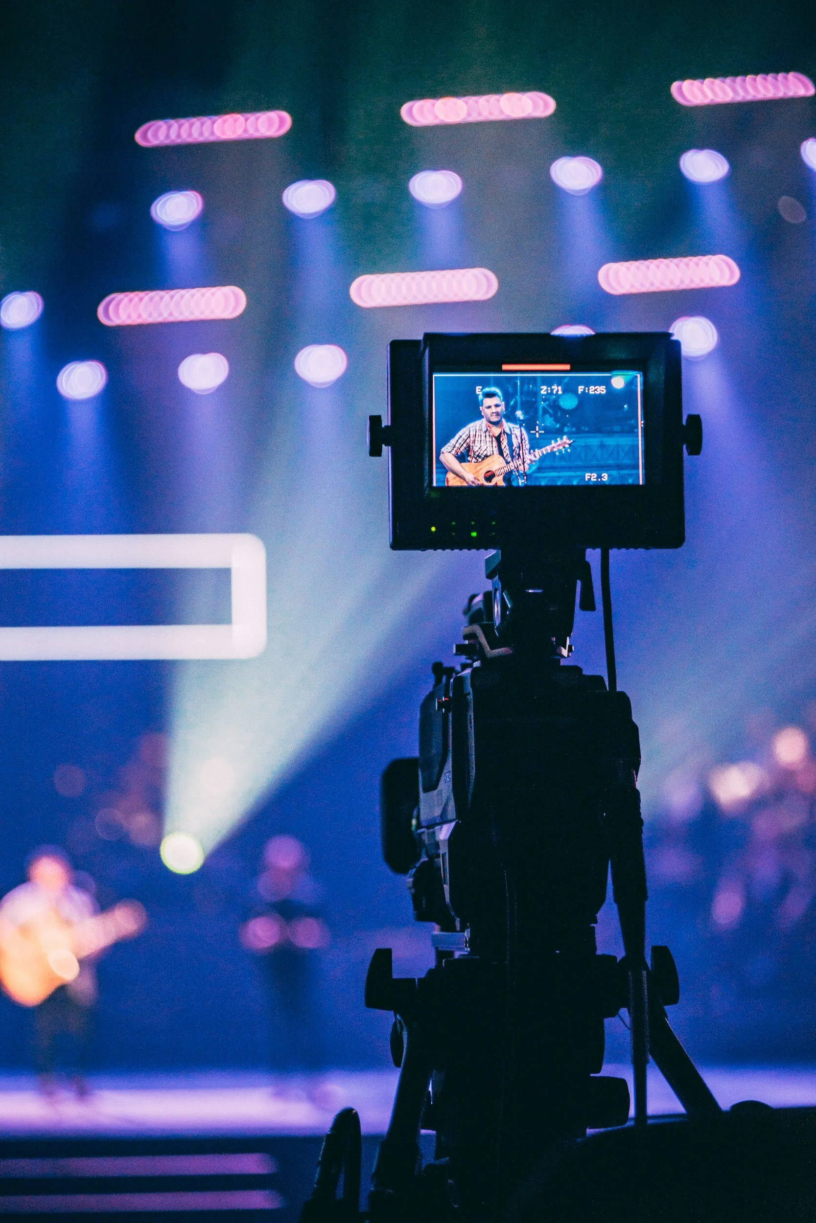 A professional video camera is recording a man playing a guitar on stage during a concert or event with colorful stage lights in the background.