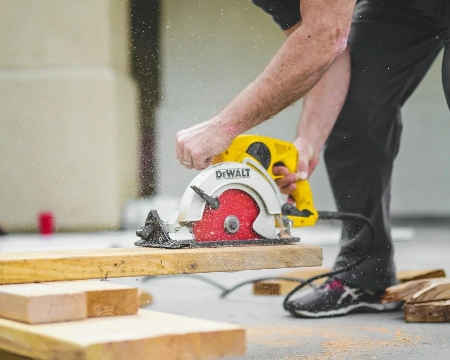A person using a yellow and red DeWalt circular saw to cut a piece of wood on a workbench, with sawdust flying in the air.