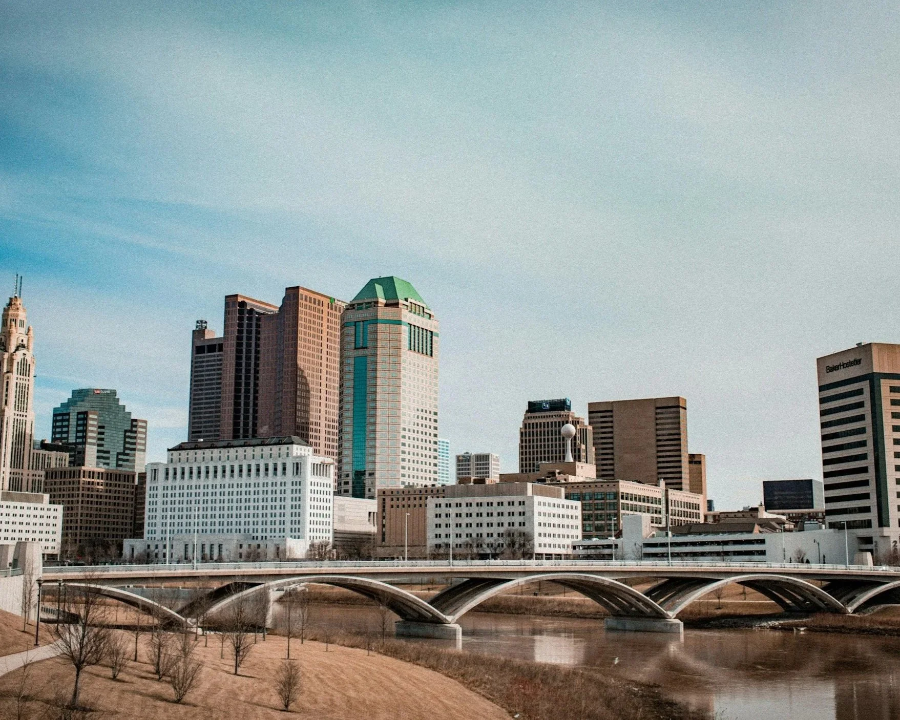 A city skyline with tall modern buildings, a bridge crossing a river, and a park with trees in the foreground, under a clear sky.