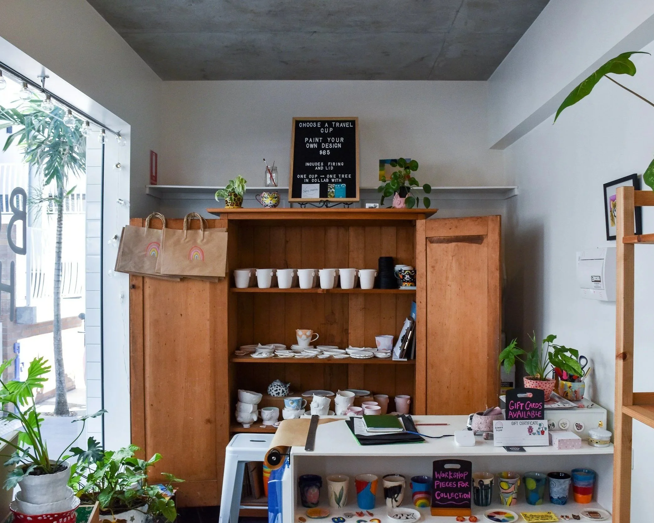 Interior of a shop with wooden shelves displaying various ceramic cups and mugs, decorated with plants and signs advertising gift cards and workshop pieces for collection.
