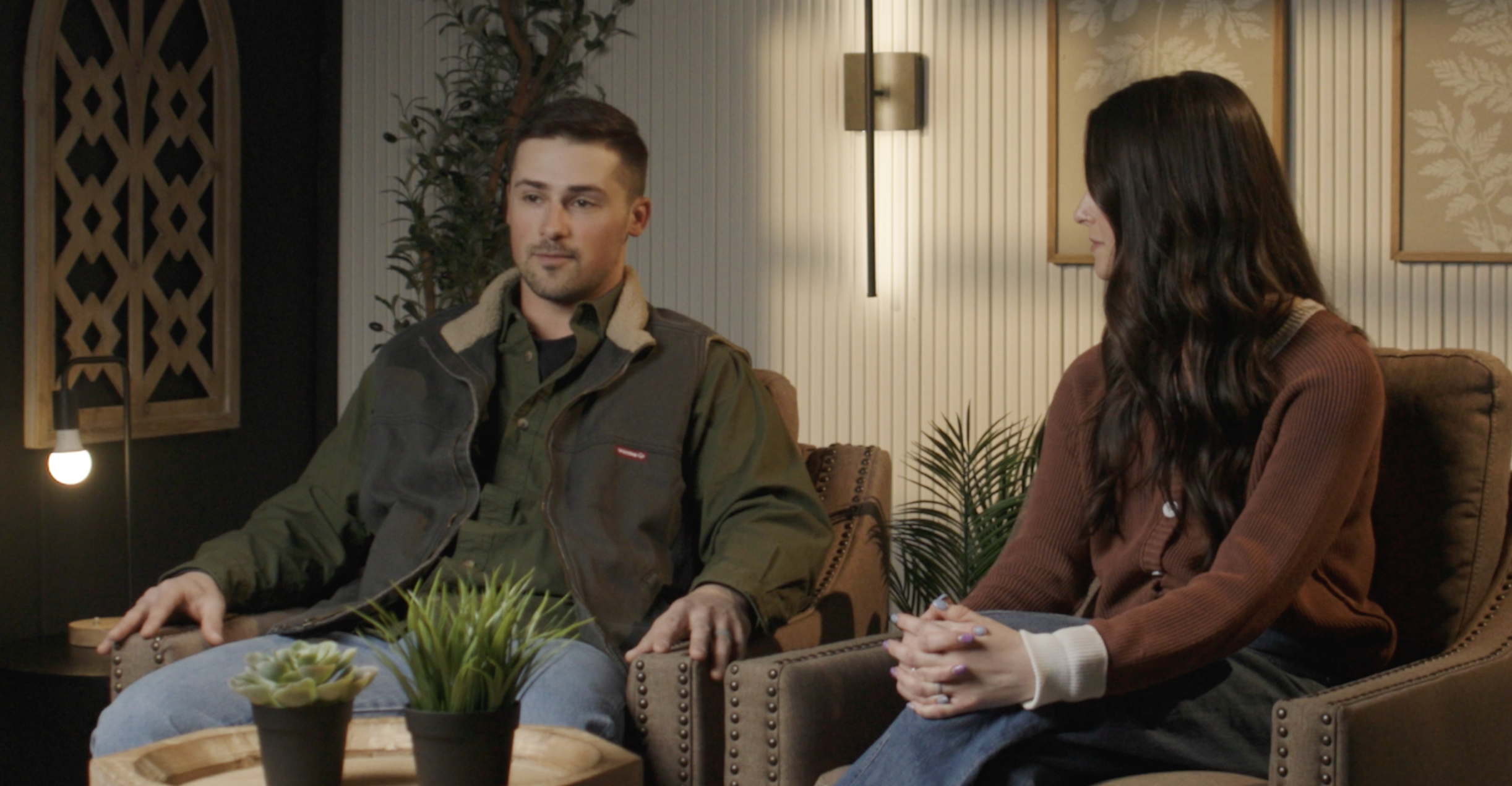 A man and a woman sitting on a brown couch in a cozy, modern room, engaged in conversation. The man has short dark hair and wears a green and black jacket, while the woman has long dark hair and wears a brown sweater. There are plants and decorative wall art behind them, with warm lighting creating a relaxed atmosphere.