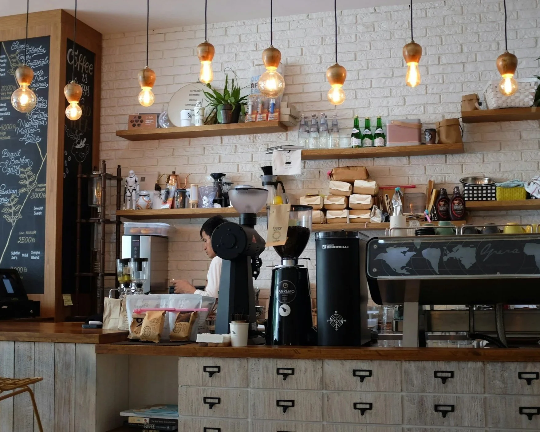 Interior of a coffee shop with a barista preparing coffee behind the counter, hanging pendant lights, shelves with bottles and plants, and a chalkboard menu.
