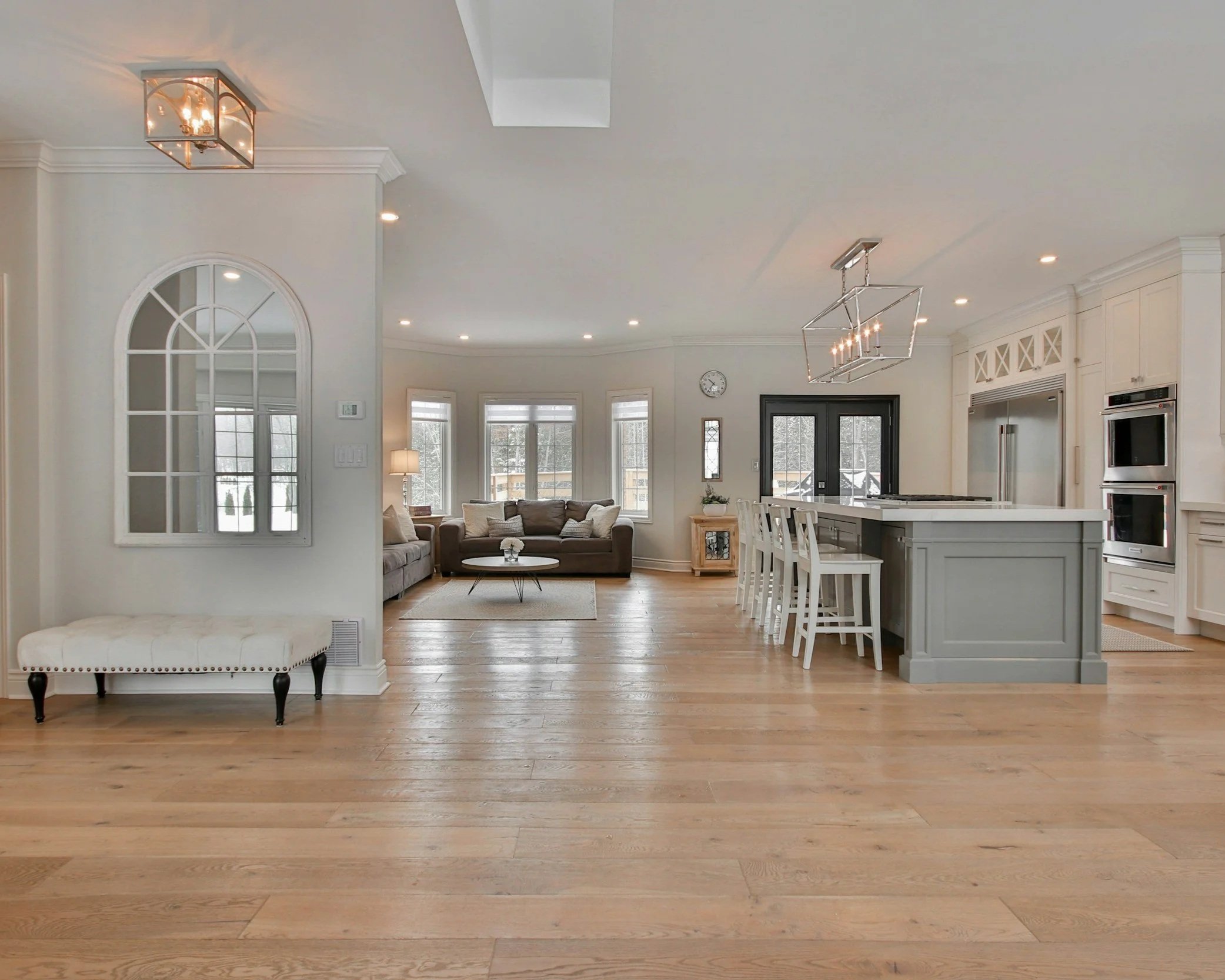 Open concept living room and kitchen with hardwood floors, white walls, large windows, and modern lighting fixtures.