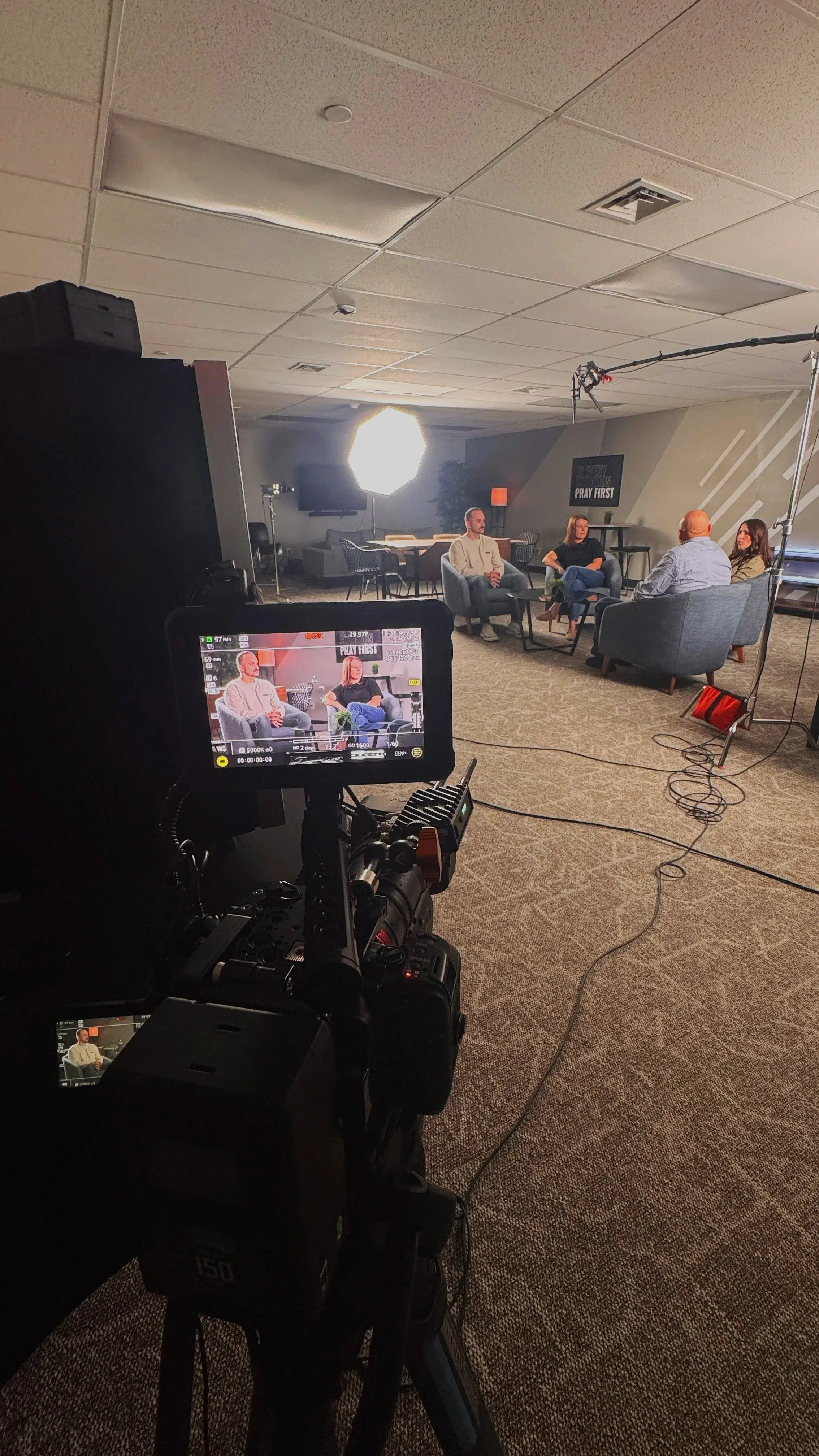 A camera and filming equipment set up in a room with four people sitting on chairs and talking, with a light source illuminating the scene. There are two table lamps, one orange and one black, along with a TV and plants in the background. A sign on the wall reads 'PRAY FIRST.'