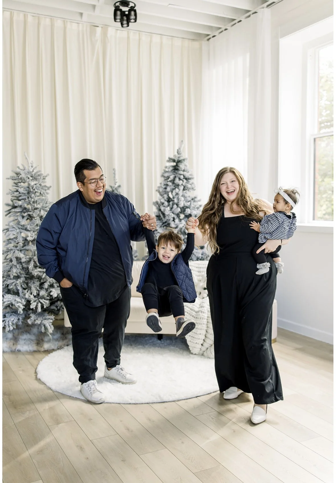 A family of four celebrating Christmas indoors, with snow-covered Christmas trees in the background. The father, mother, and two children are smiling and holding hands, with one child jumping in the air.