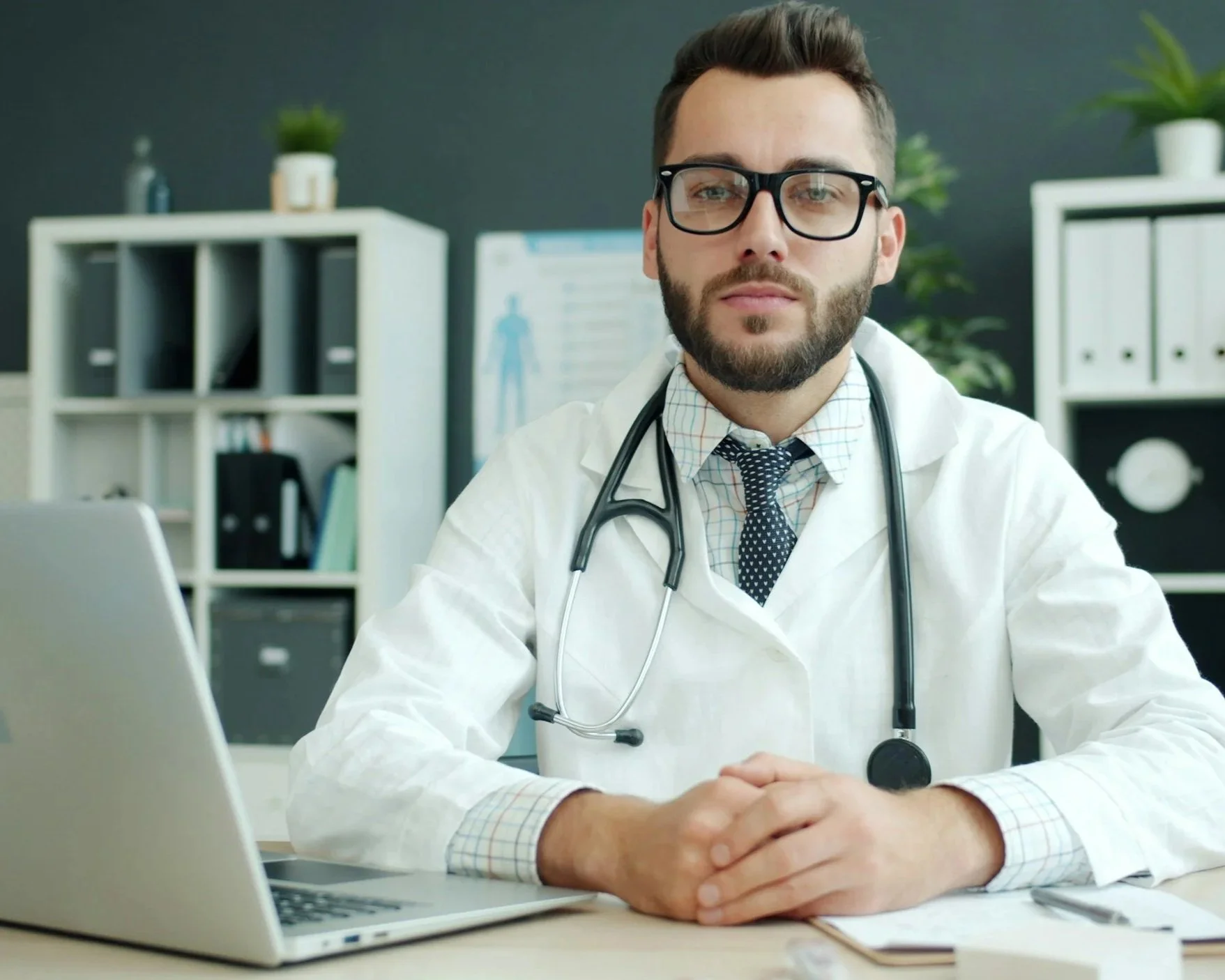 A male doctor wearing glasses, a white lab coat, and a stethoscope around his neck, sitting at a desk with a laptop, in an office with shelves and plants in the background.