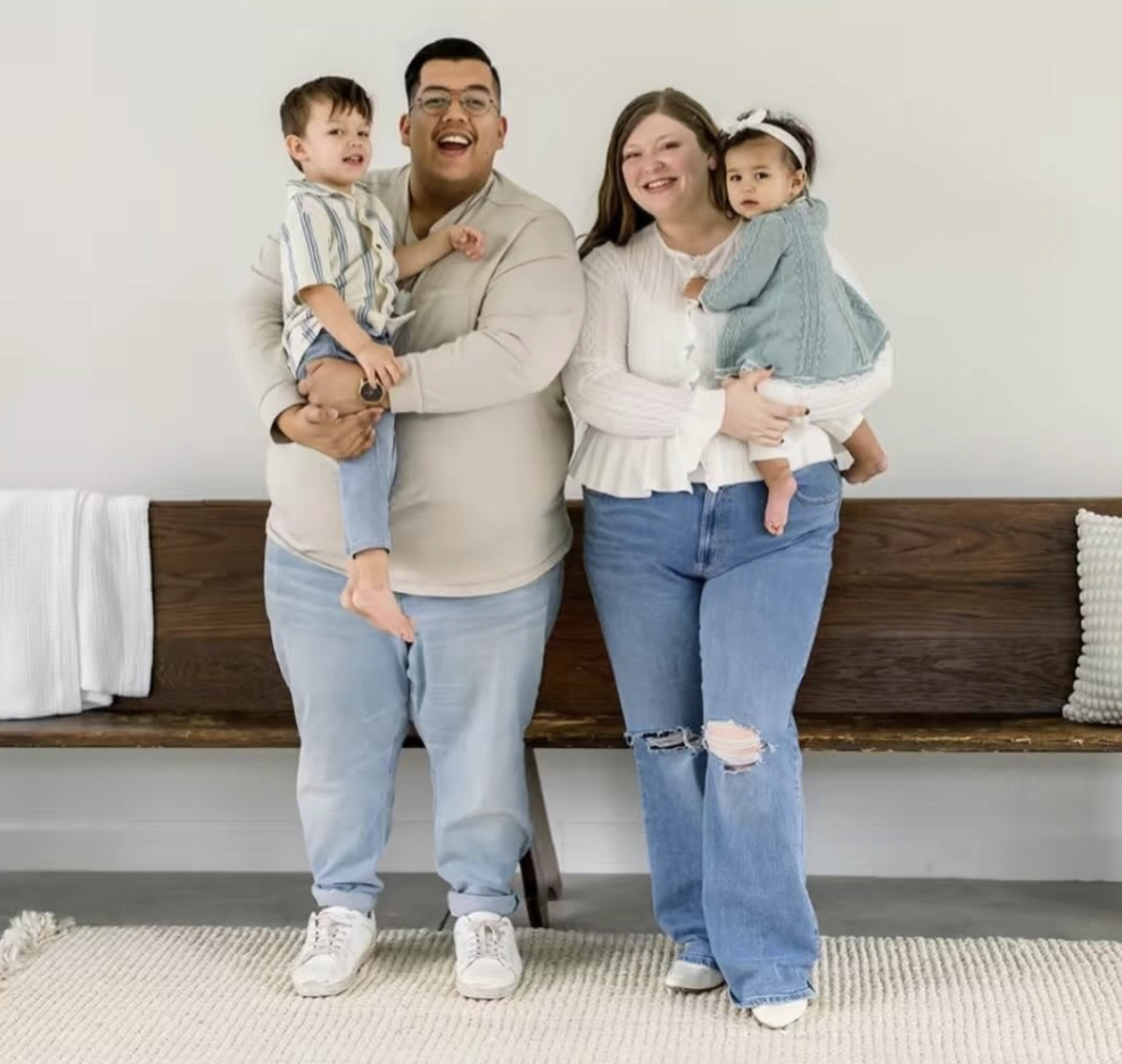 A smiling family of four standing in front of a white wall. The father holds a young boy, and the mother holds a young girl. The family appears happy and is casually dressed.