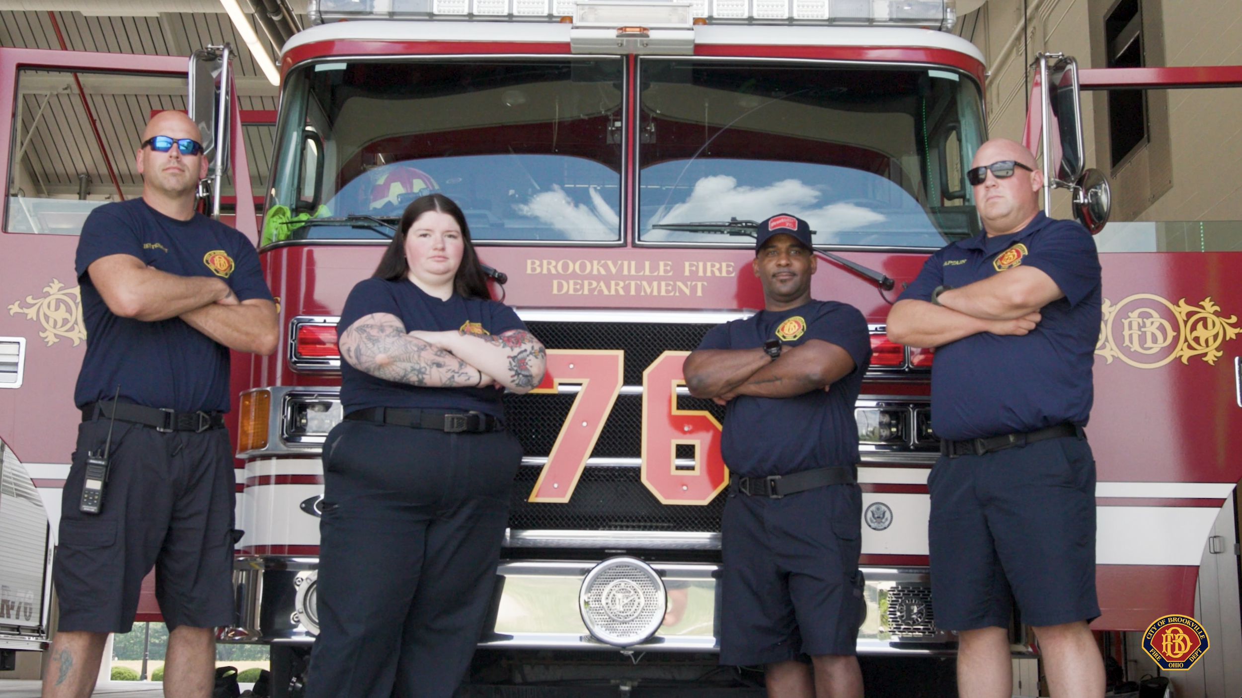 Four firefighters stand in front of a red fire truck with 'Brookville Fire Department' written on it. They have crossed arms and are wearing navy blue shirts with fire department patches, with two wearing sunglasses. One firefighter has tattoos on her arms.