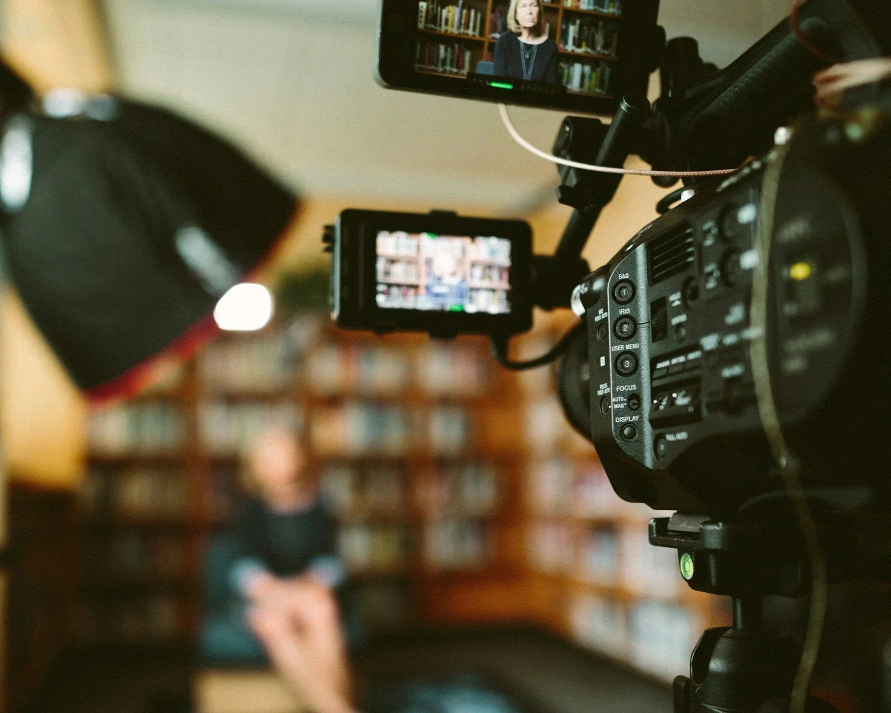A professional video camera filming a woman speaking in a library. The shot shows the camera's viewfinder displaying the woman in front of bookshelves.