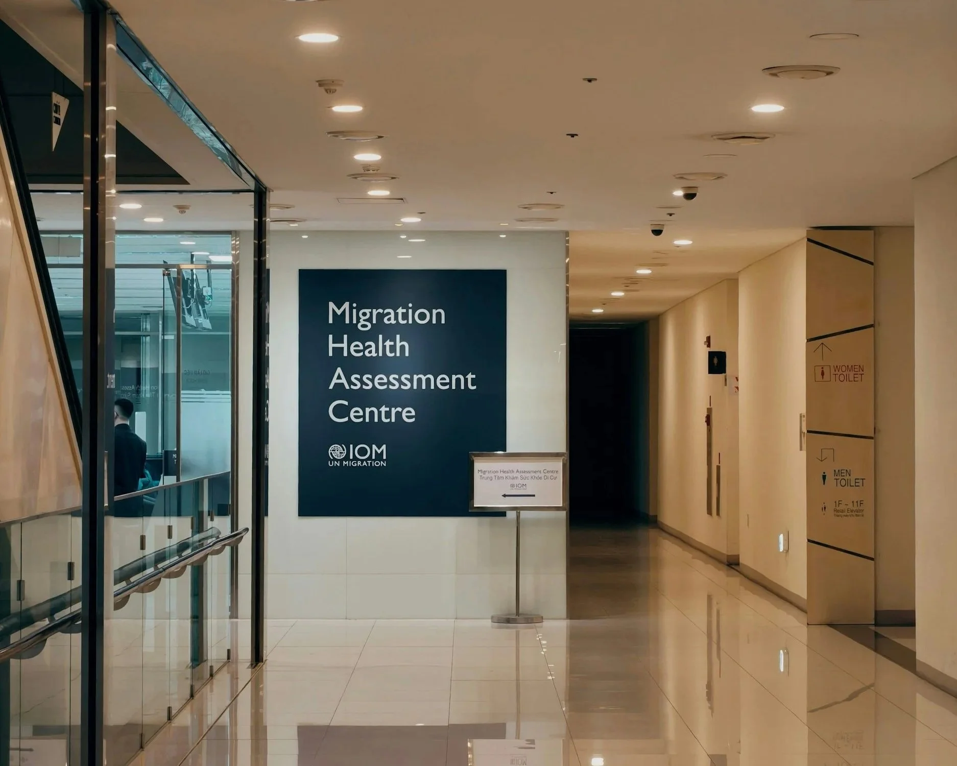 Interior corridor of a building with signs for a Migration Health Assessment Centre and restrooms. Bright lighting, glossy beige tiles, and elevator doors are visible.
