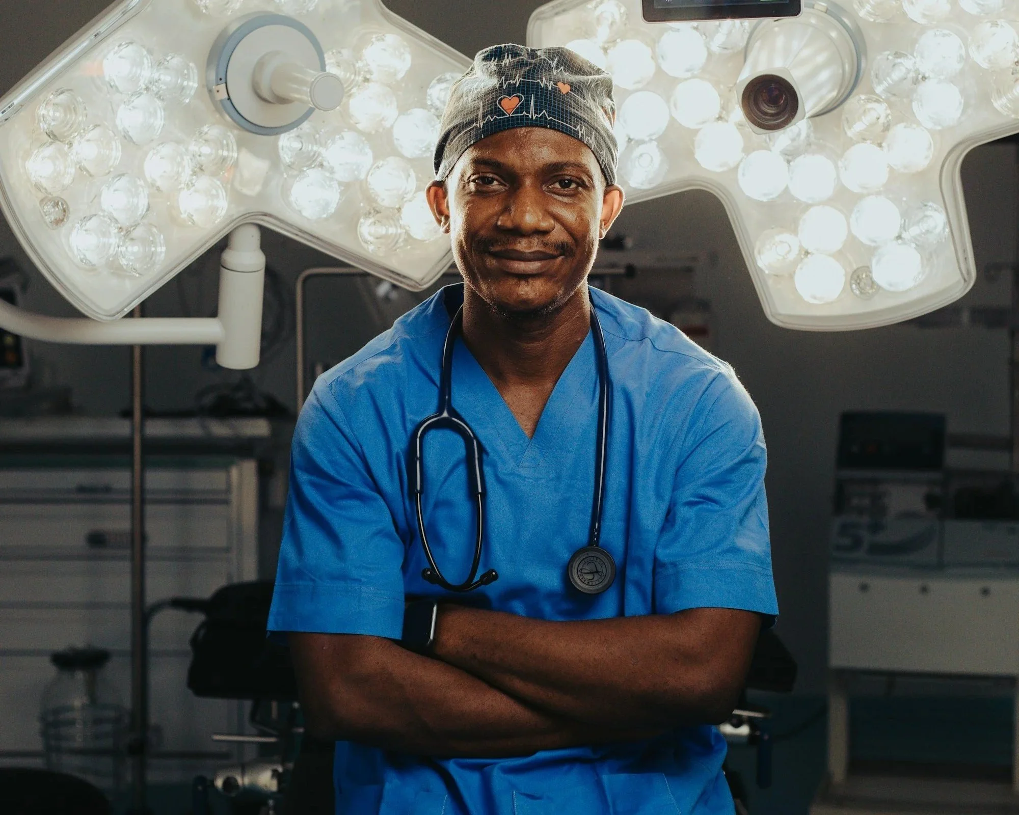 A male doctor wearing blue scrubs, a gray medical cap with hearts and heartbeat lines, and a stethoscope around his neck, standing in an operating room with surgical lights overhead.
