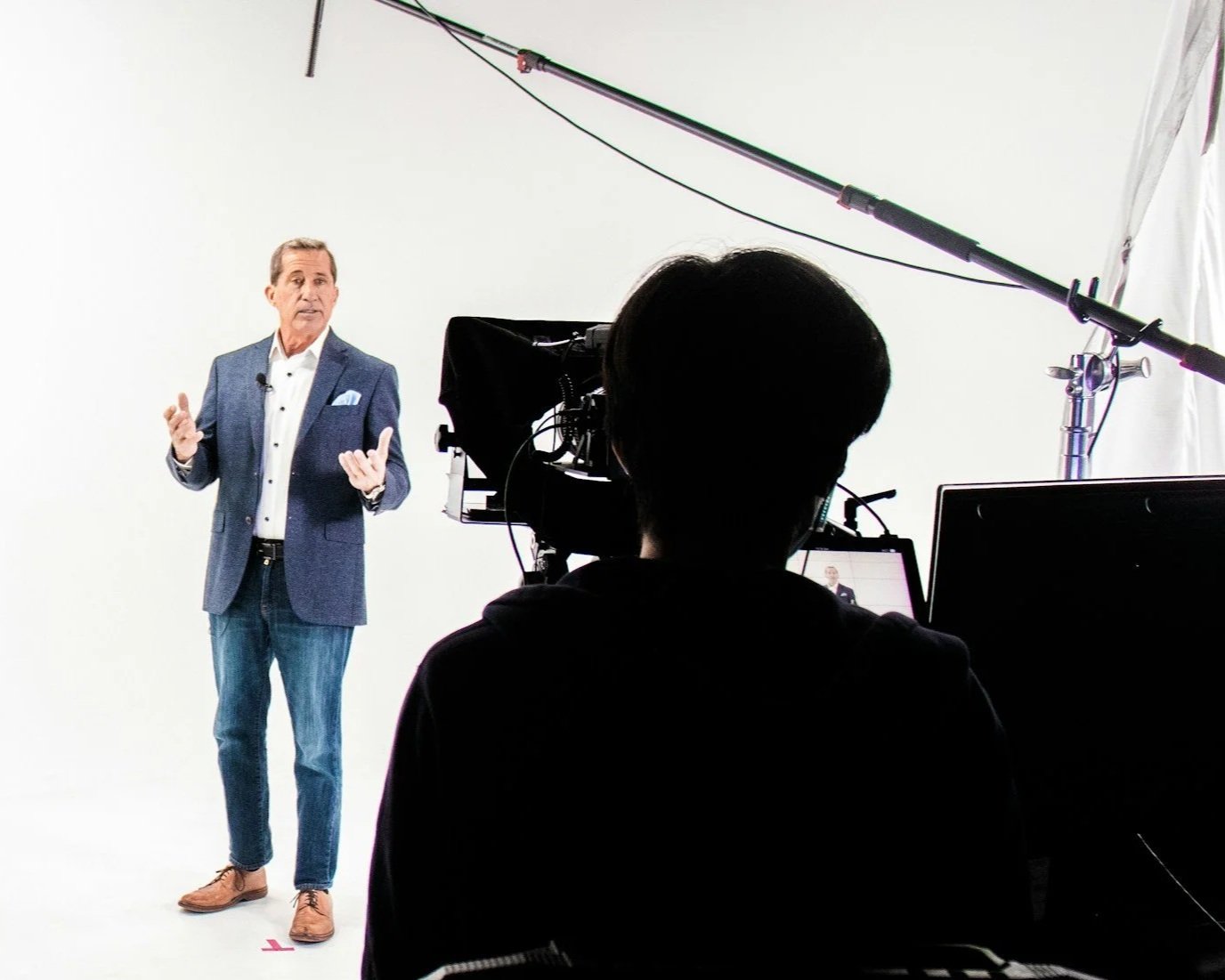 A man wearing a blue blazer and jeans is speaking in front of a camera in a white studio, with a crew member operating the camera.