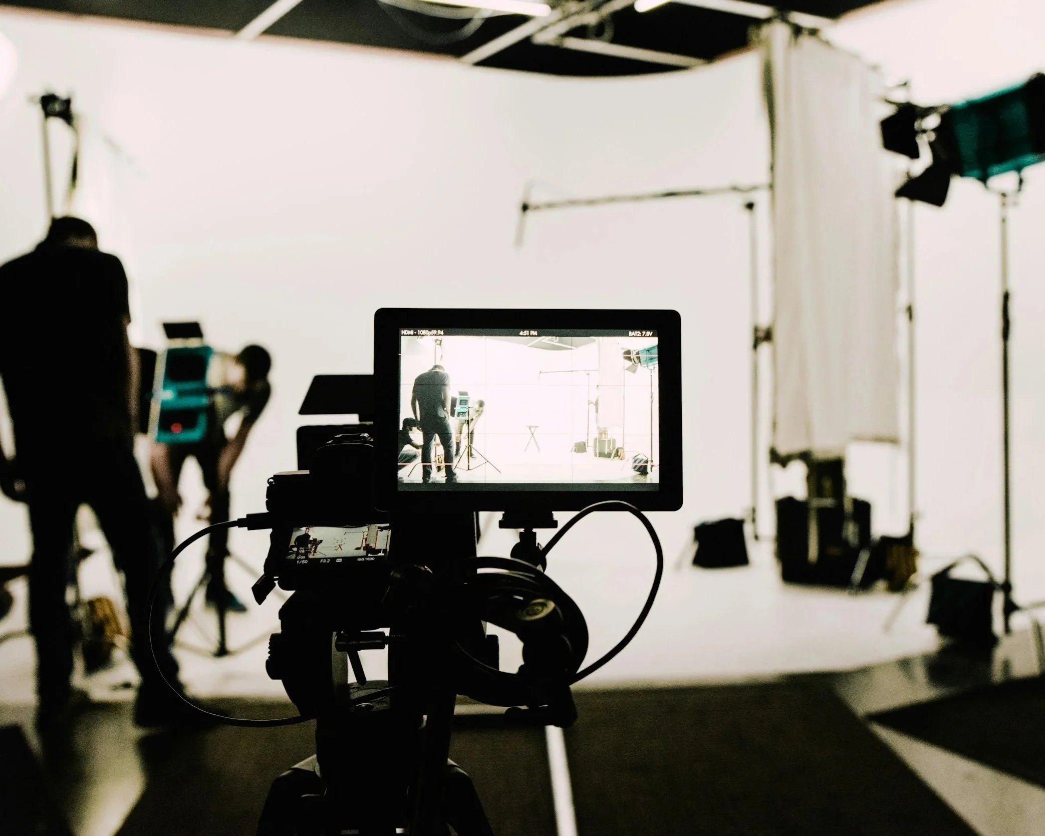 A film or photography studio with a camera on a tripod facing a white backdrop. A monitor in the foreground displays a scene being filmed, including a person standing near equipment. Studio lights and equipment are arranged around the set.