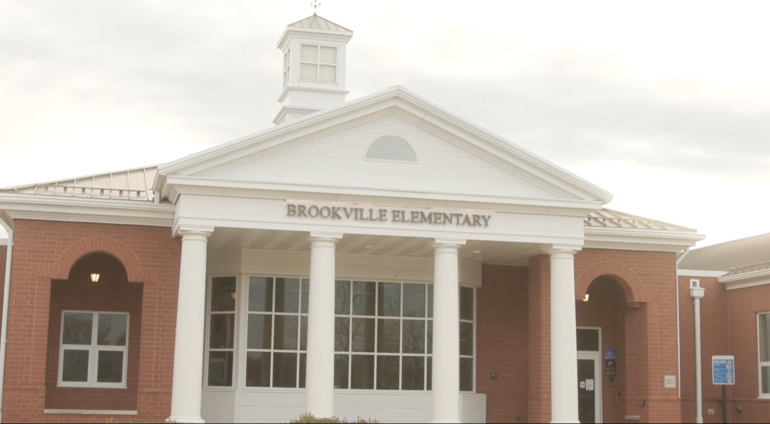 Front view of Brookville Elementary school building with white columns and brick facade, topped with a white triangular pediment and a small tower on the roof, under cloudy sky.