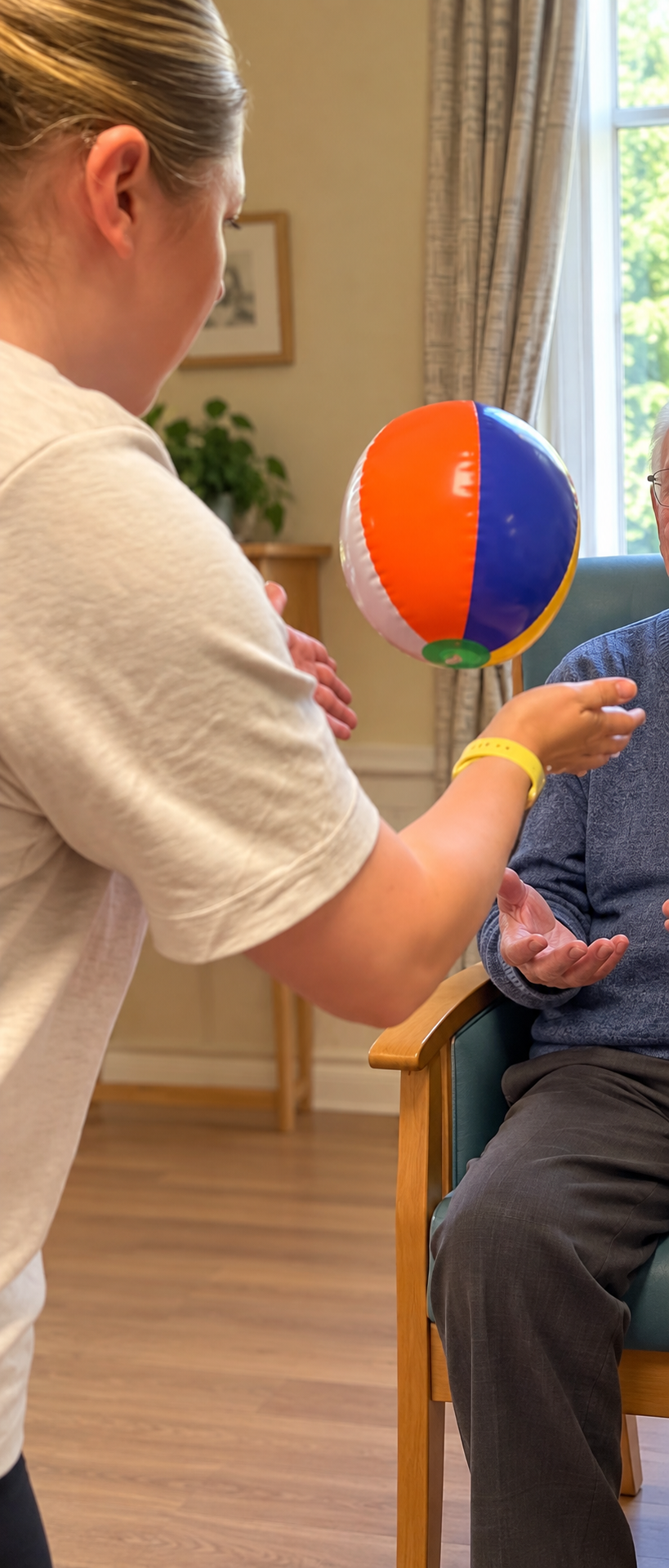 A young woman in a beige t-shirt playing with a beach ball with a senior person seated in a chair, indoors, with a window showing trees outside.