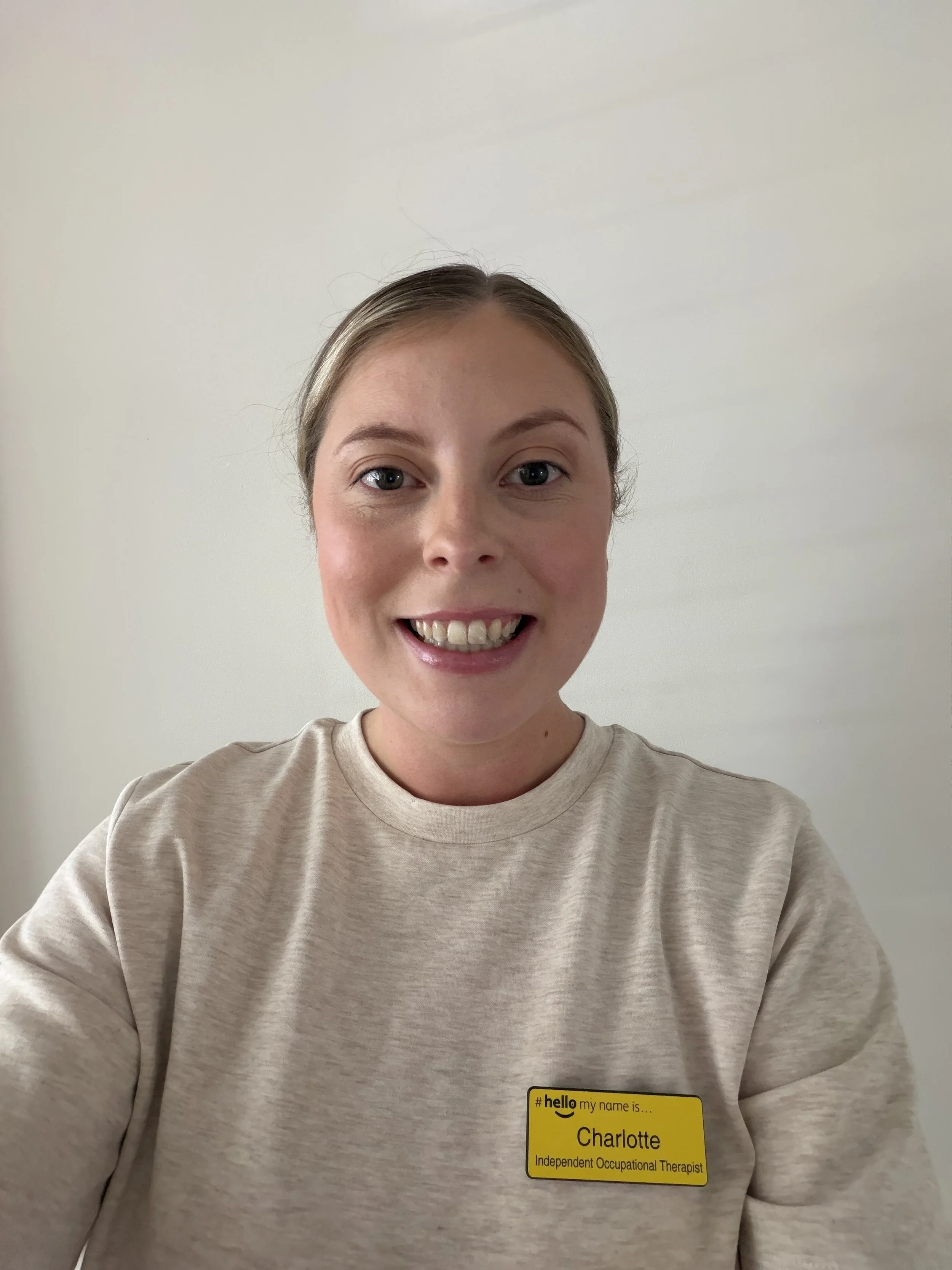 A woman with blonde hair and blue eyes smiling, wearing a beige shirt and a yellow name badge with black text that reads 'Charlotte, Independent Occupational Therapist,' against a plain light-colored wall.