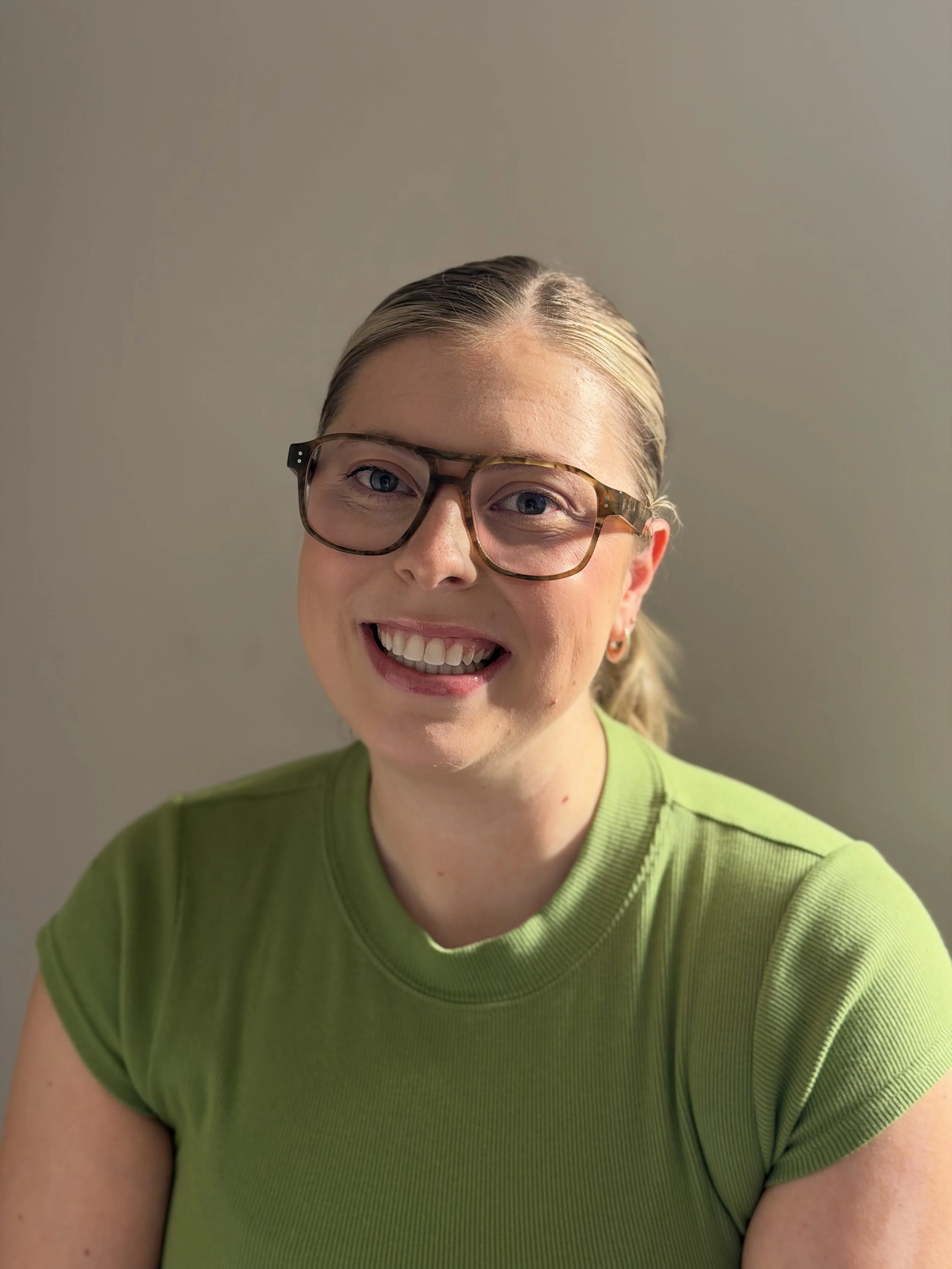 A woman with blonde hair pulled back, wearing thick-rimmed glasses and a bright green t-shirt, smiling in front of a plain, light-colored background.