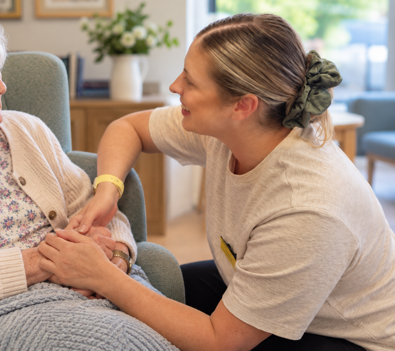 A caregiver smiling and holding hands of an elderly woman, engaging in a caring interaction in a cozy, well-lit home environment.