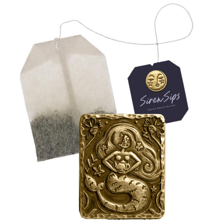 A tea bag with black tea leaves, a decorative golden Mermaid-shaped soap or ornament with engraved details, and a navy blue tag with a gold moon face button attached, labeled "Siren Sips Ocean Dream Blend".