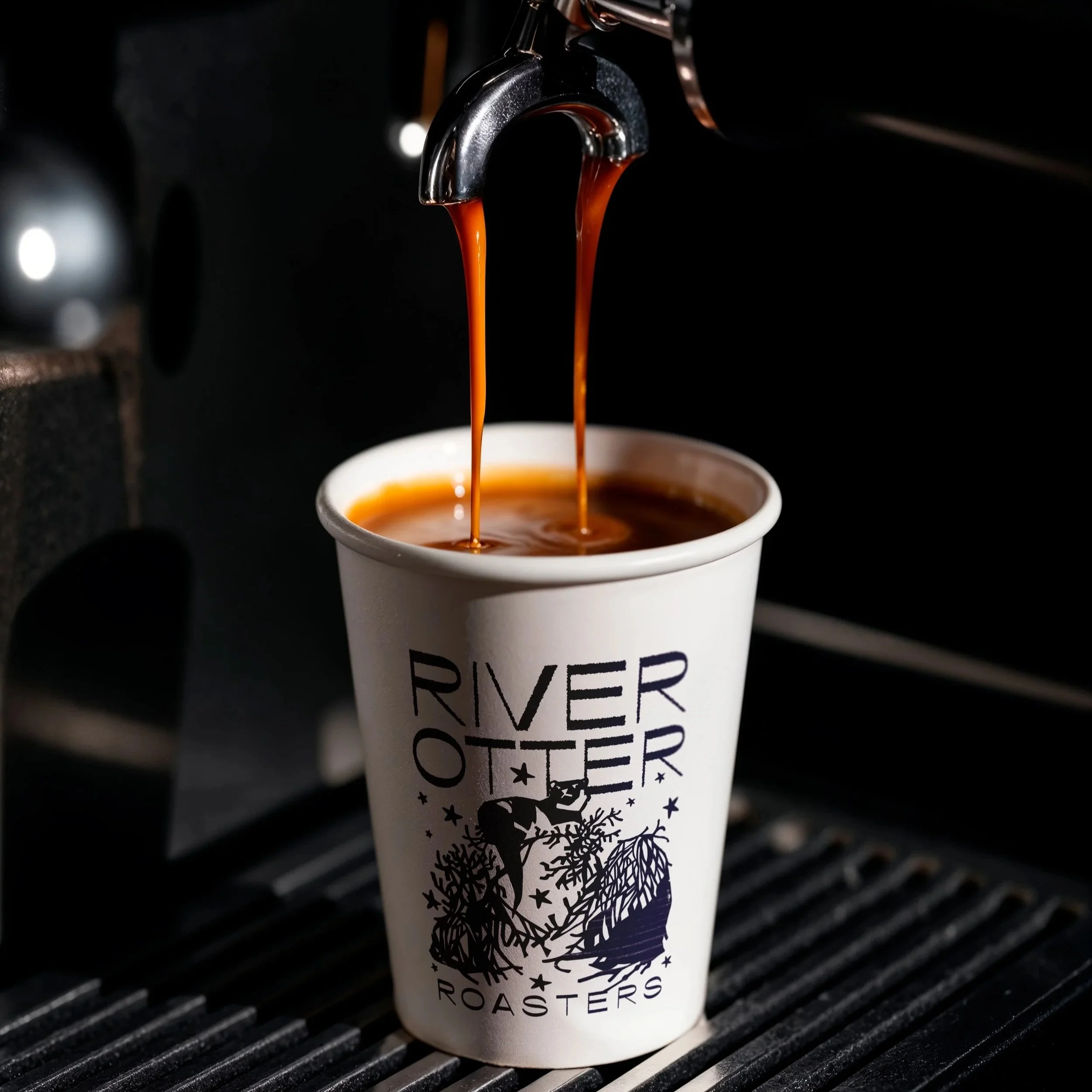 Freshly brewed coffee being poured into a white disposable cup with a 'River Otter Roasters' logo, from a coffee machine, on a black coffee drip tray.