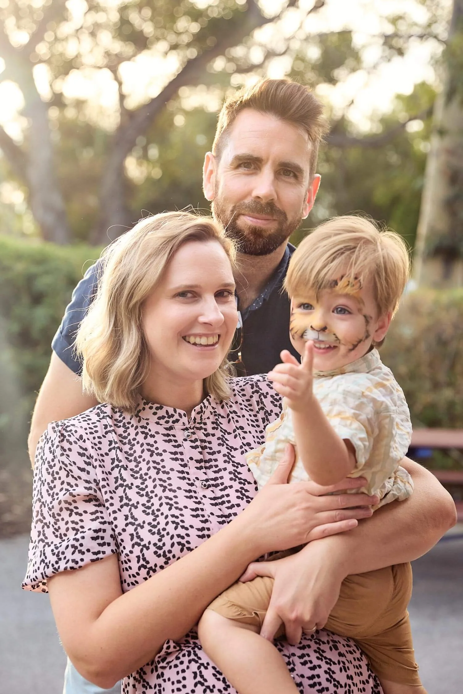 A family of three posing outdoors in a park with trees in the background. The woman is holding a young boy with face paint of a tiger, and a man stands behind them smiling.