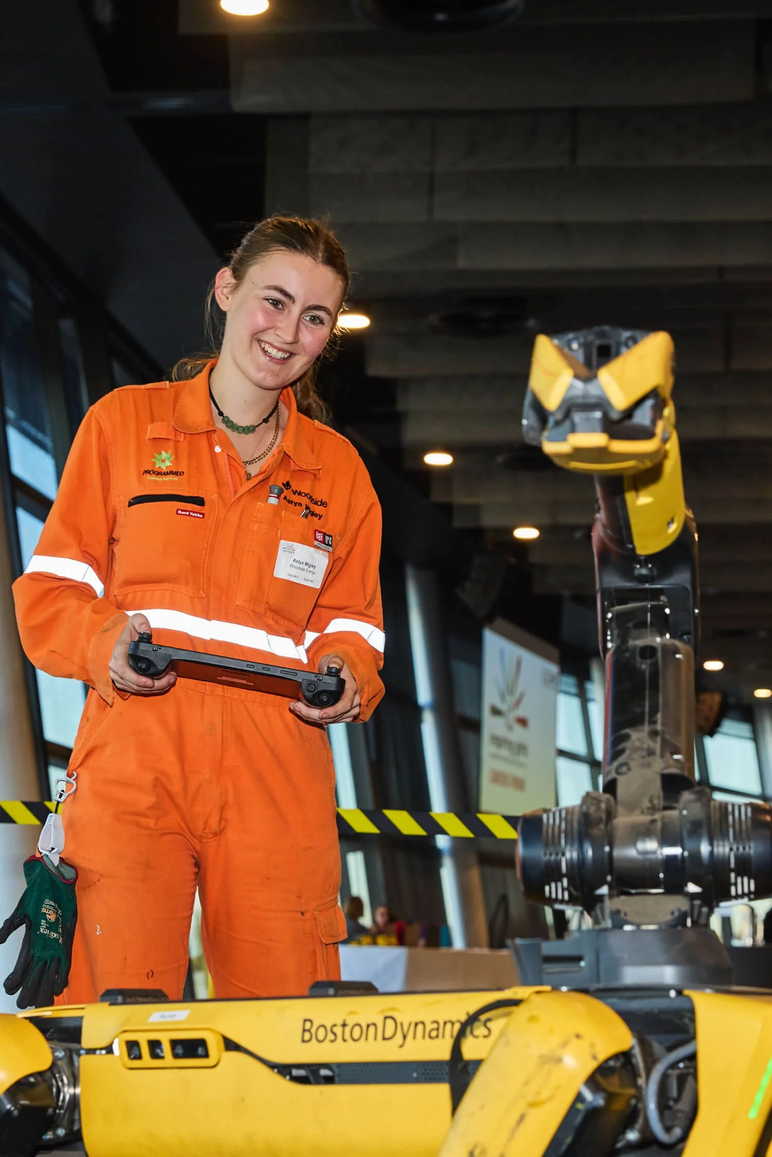 A woman in orange safety gear smiling while operating a remote control with a yellow robot in an indoor setting.