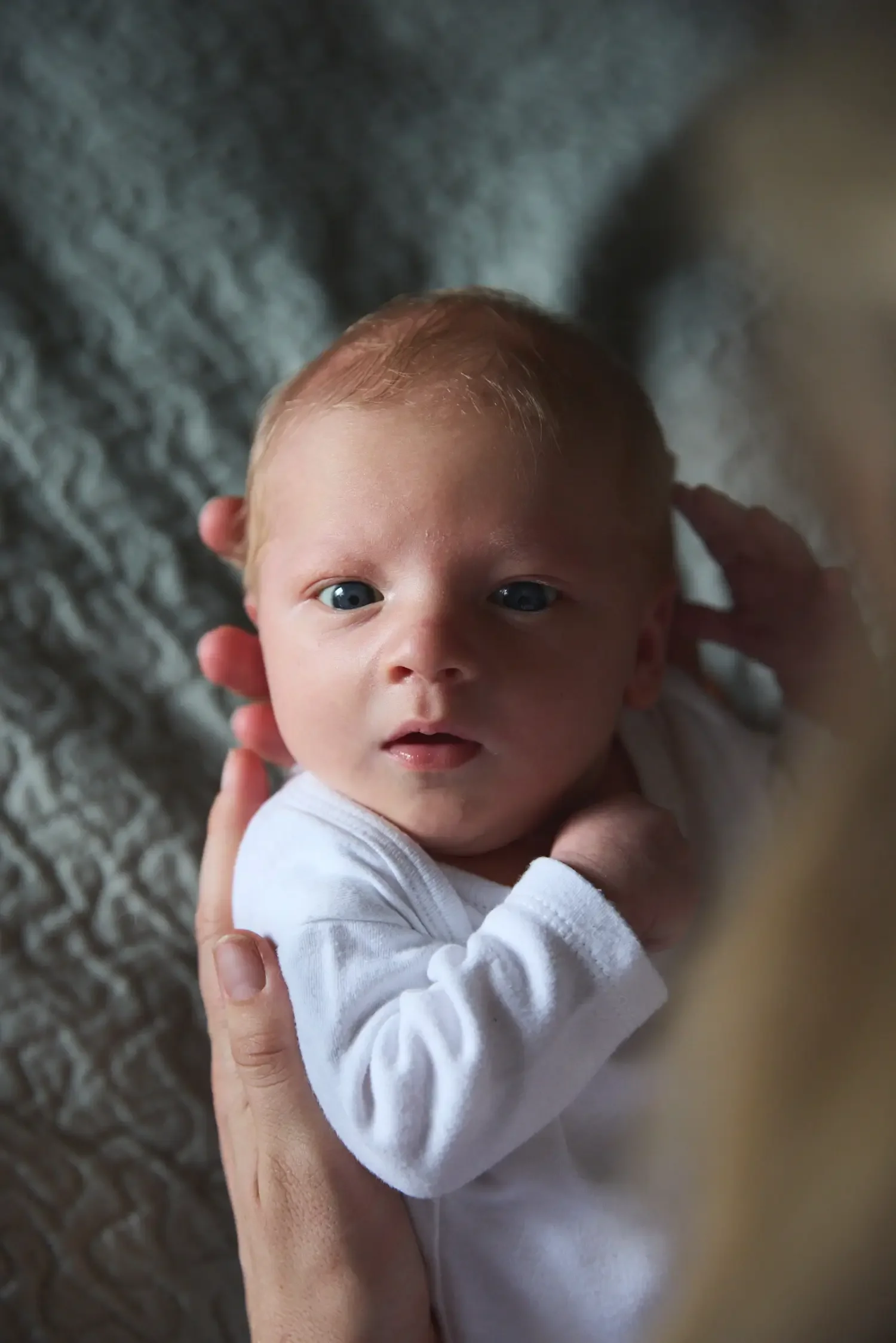 Close-up of a baby with blonde hair and blue eyes being gently held and supported by an adult's hand, wearing a white shirt, against a textured gray background.