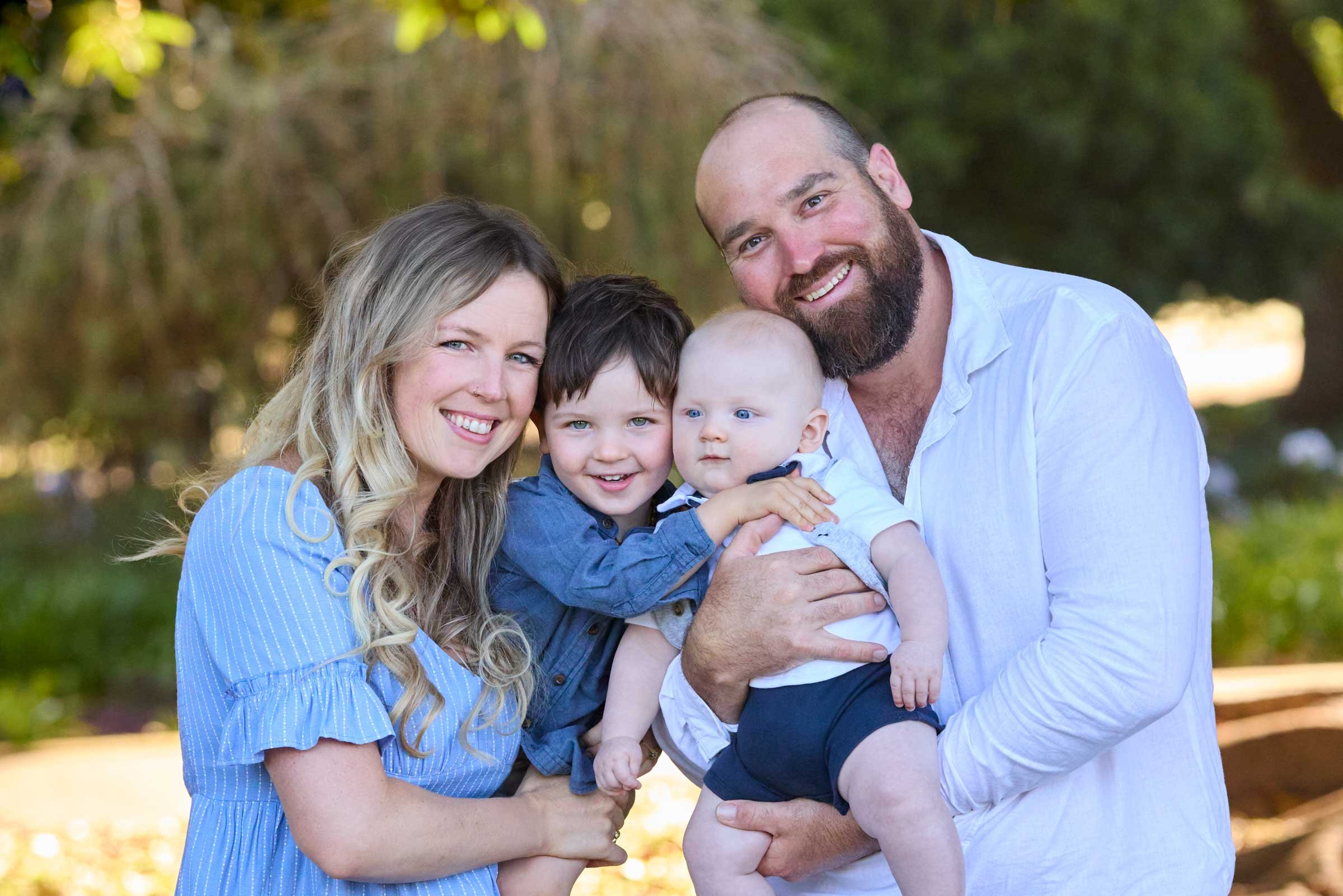 Smiling family of four outdoors, with mother, father, and two young children, surrounded by greenery and sunlight.