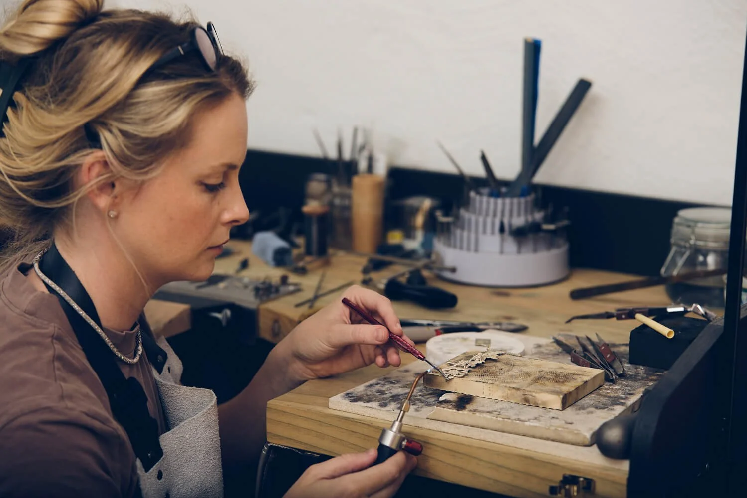 A woman working at a jewelry soldering station, holding a soldering iron and a piece of metal on a workbench filled with tools and equipment.
