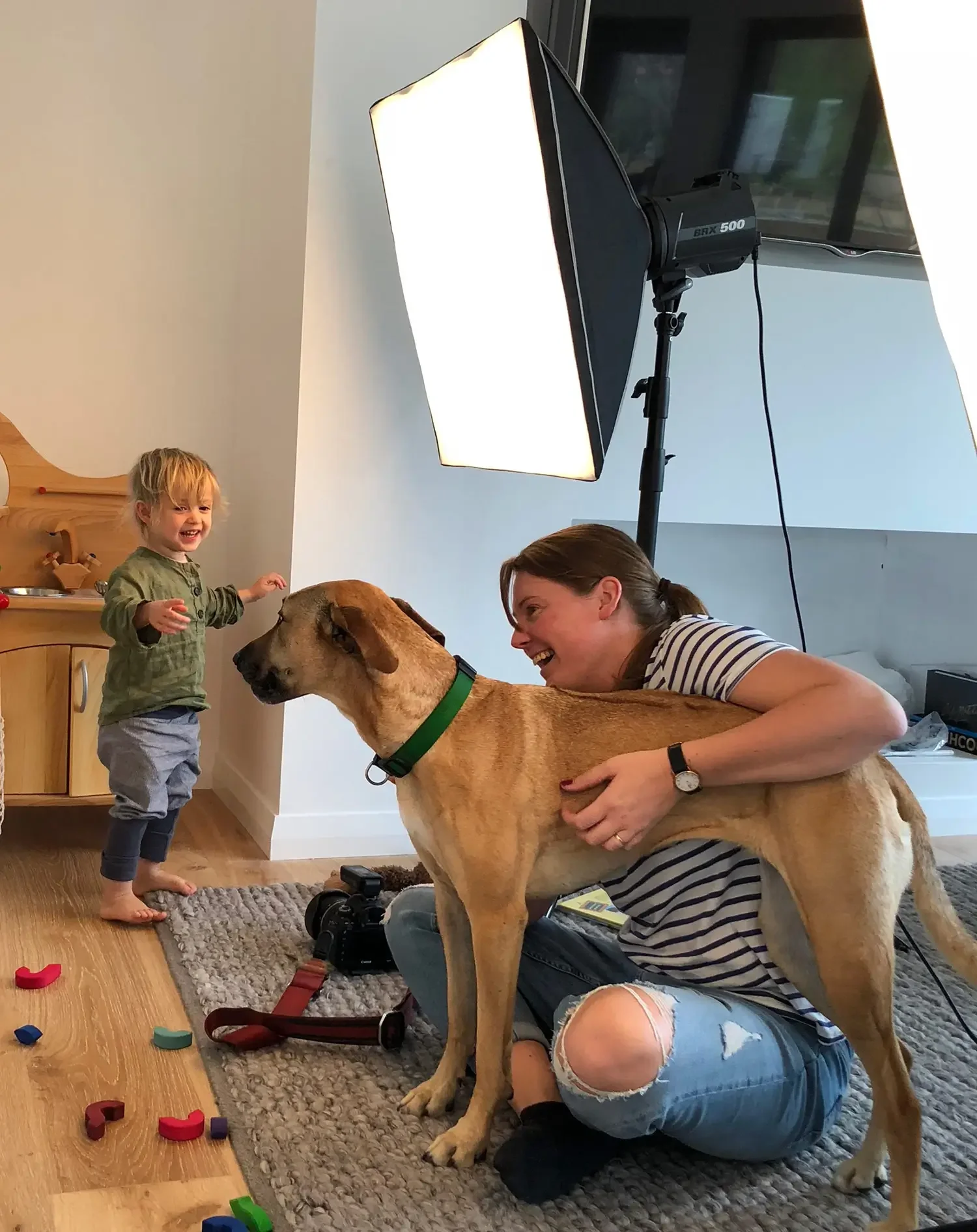 A woman and a child with a dog in a living room. The woman is sitting on the floor, smiling, with her arm around the dog. The child stands and smiles at the woman. Toys are scattered on a rug and a large studio light is set up nearby.